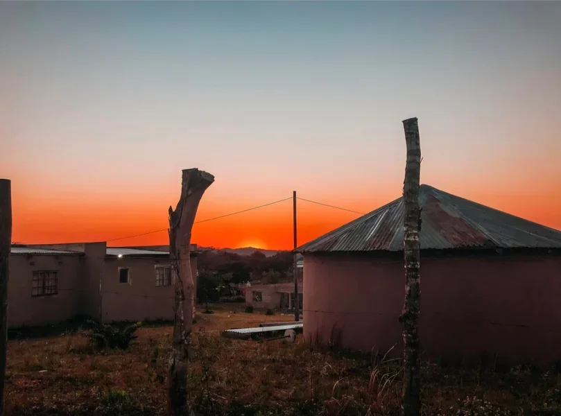 A sunset over a rural area with a pink, round building with a corrugated metal roof in the foreground, surrounded by weathered wooden posts and dry grass.