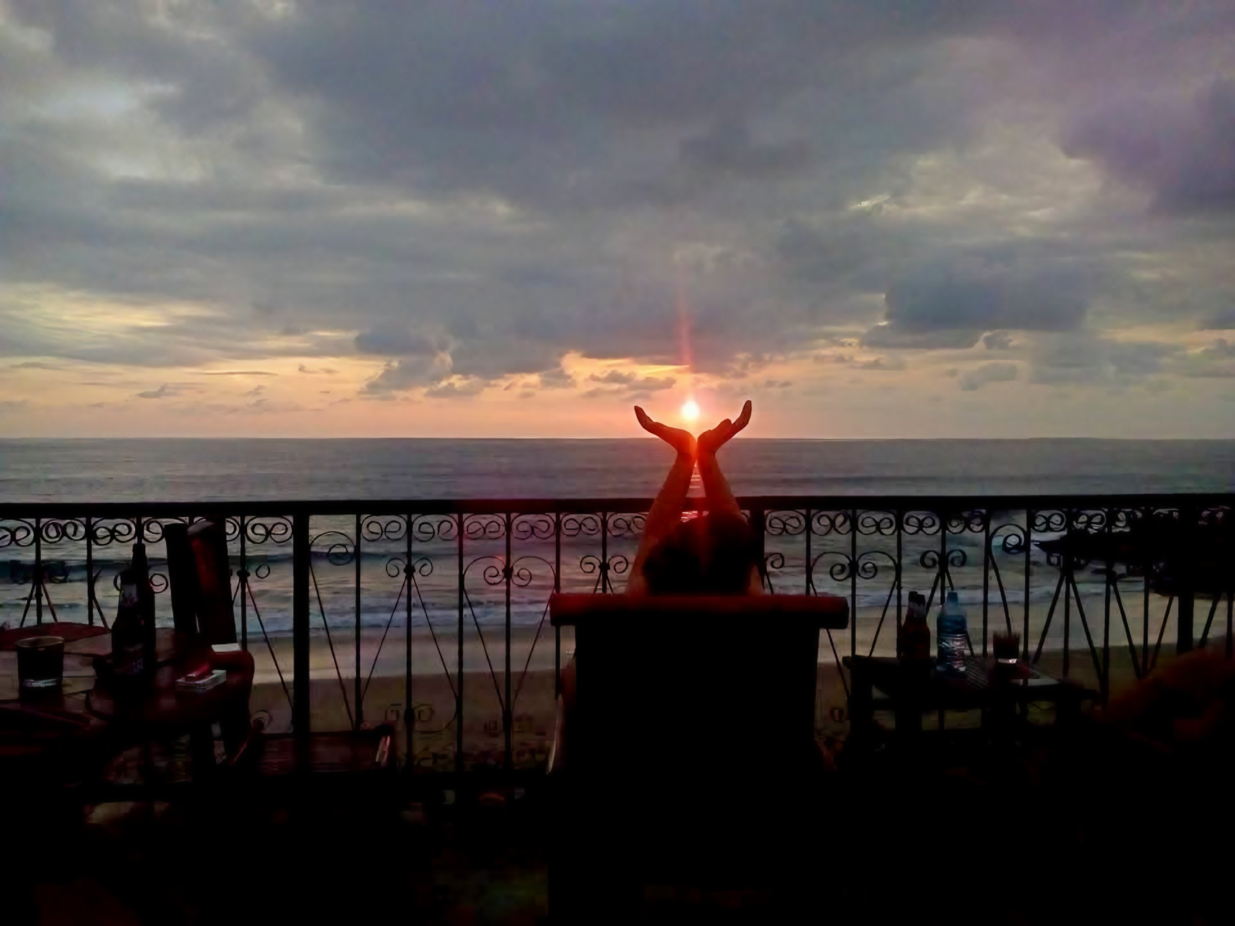 A person holding their hands up in front of the sun at sunset over the ocean, with cloudy skies above and a decorative railing in the foreground.
