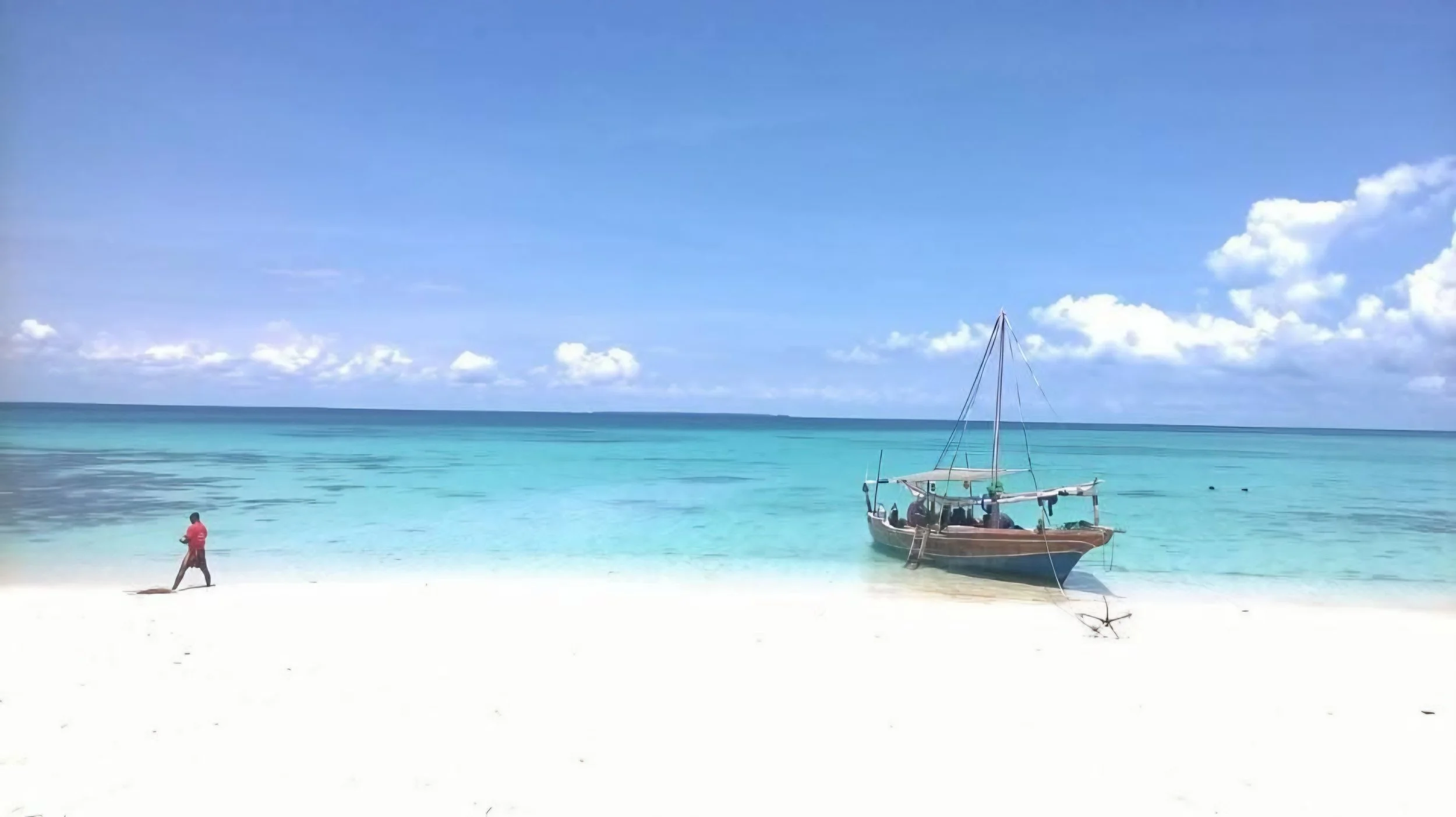 A person in a red shirt and shorts walks along the white sandy beach near clear turquoise water, with a small sailboat anchored nearby under a bright blue sky with scattered clouds.