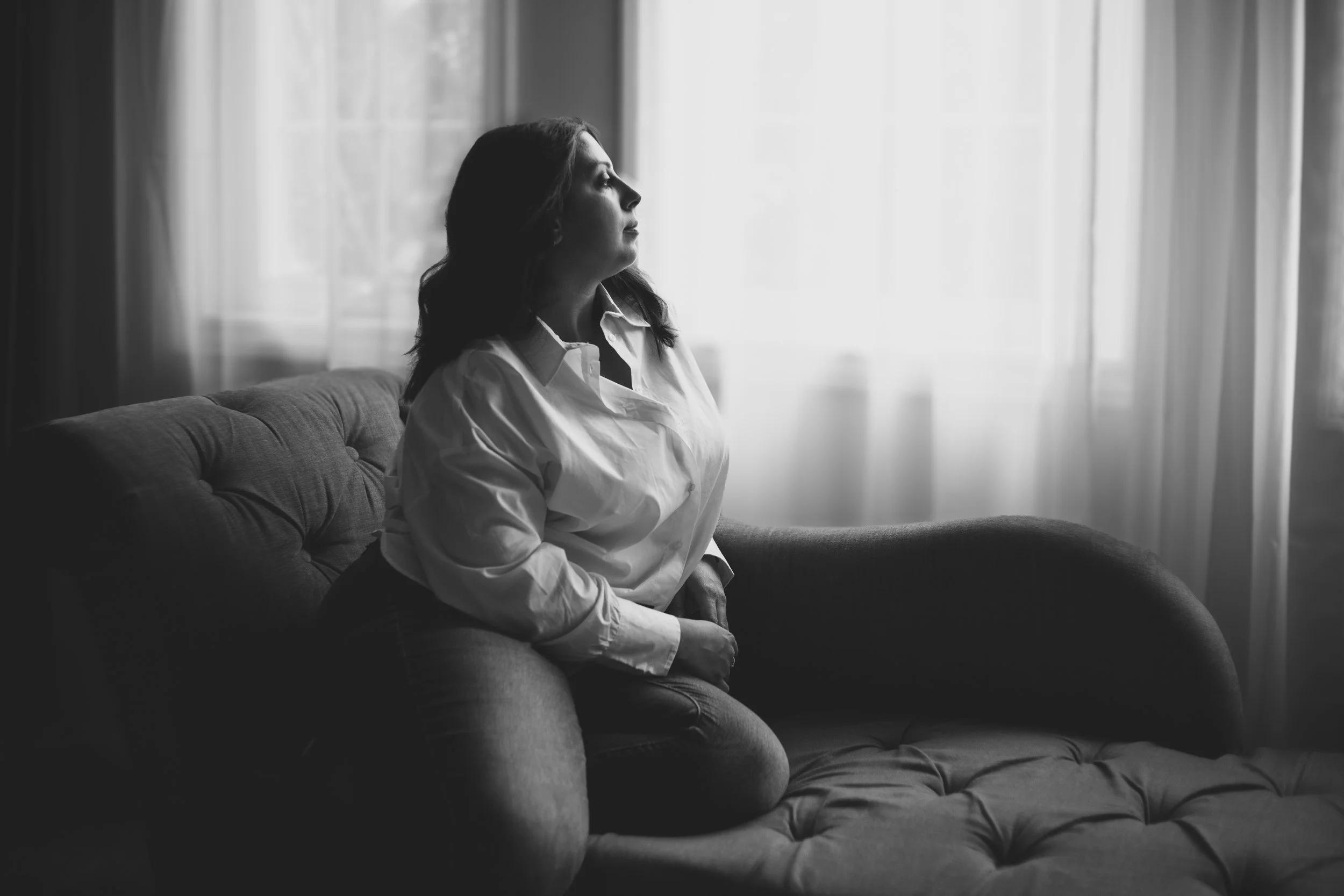 A woman with dark hair, wearing a white shirt, sitting on a couch and looking out a window with curtains. The image is in black and white.