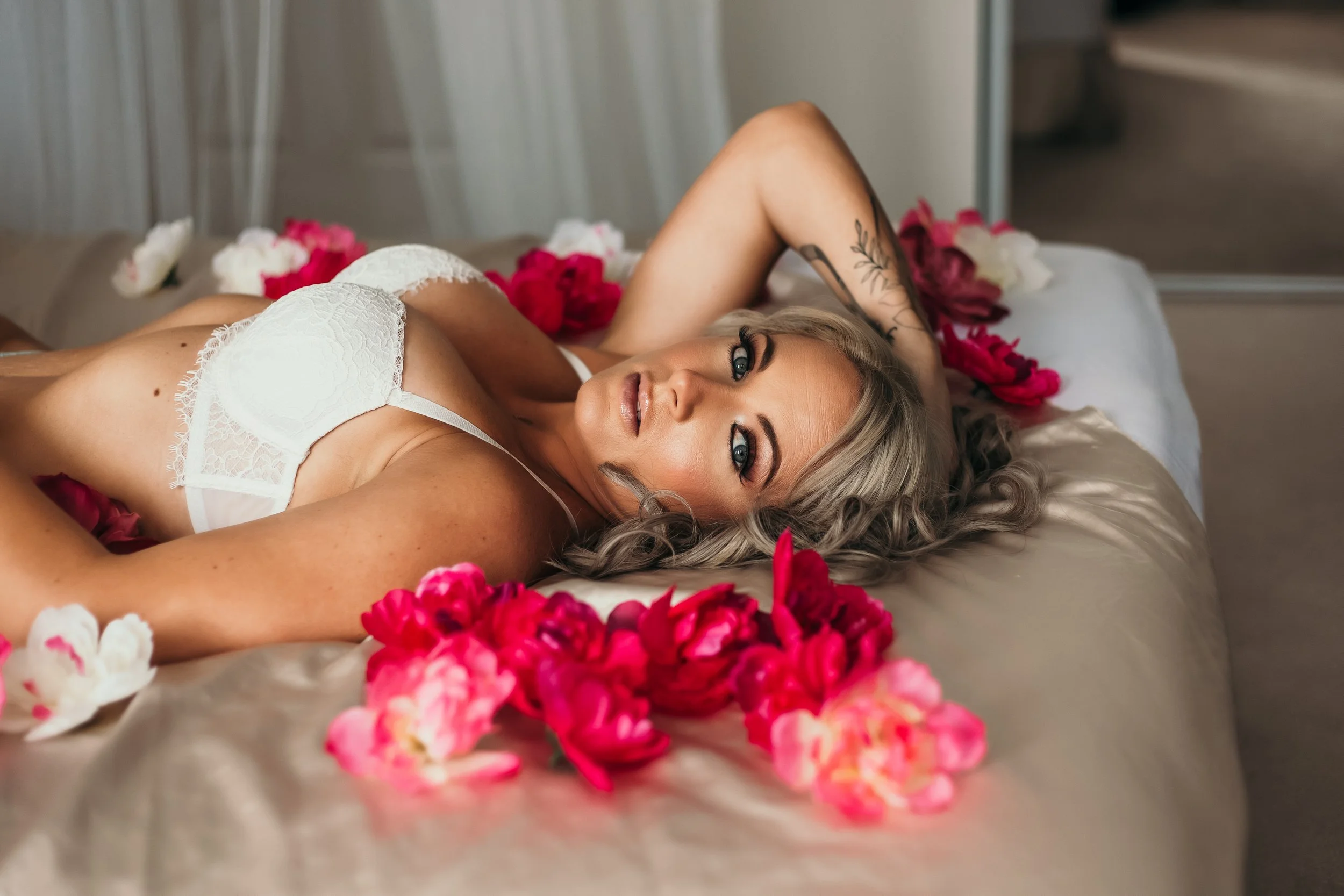 A woman with blonde curly hair lying on a bed surrounded by pink and white flowers, wearing white lace lingerie, looking at the camera.