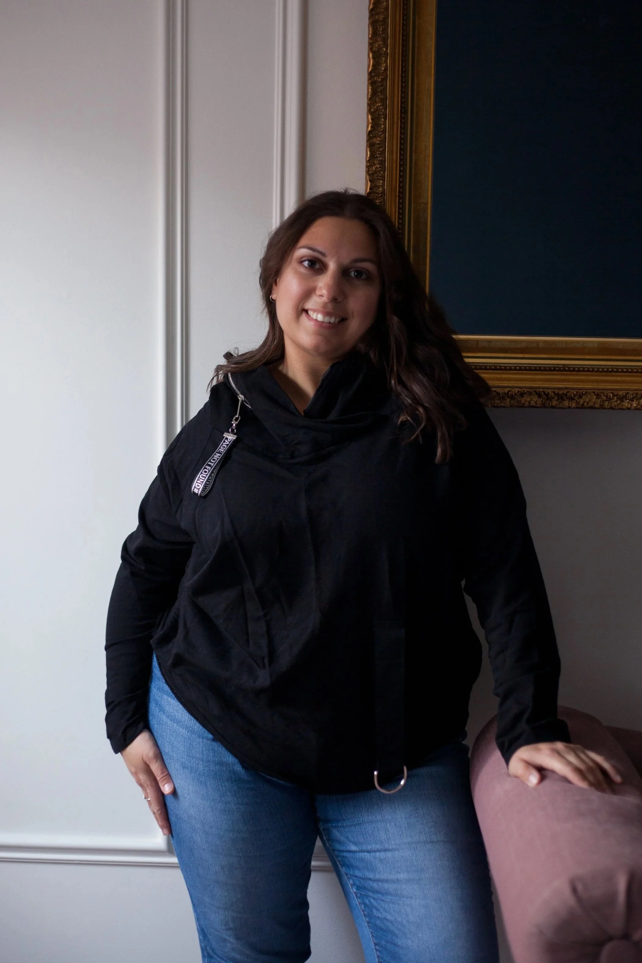A woman with brown hair, wearing a black jacket and blue jeans, standing indoors in front of a wall with decorative molding and a framed blue display, smiling at the camera.