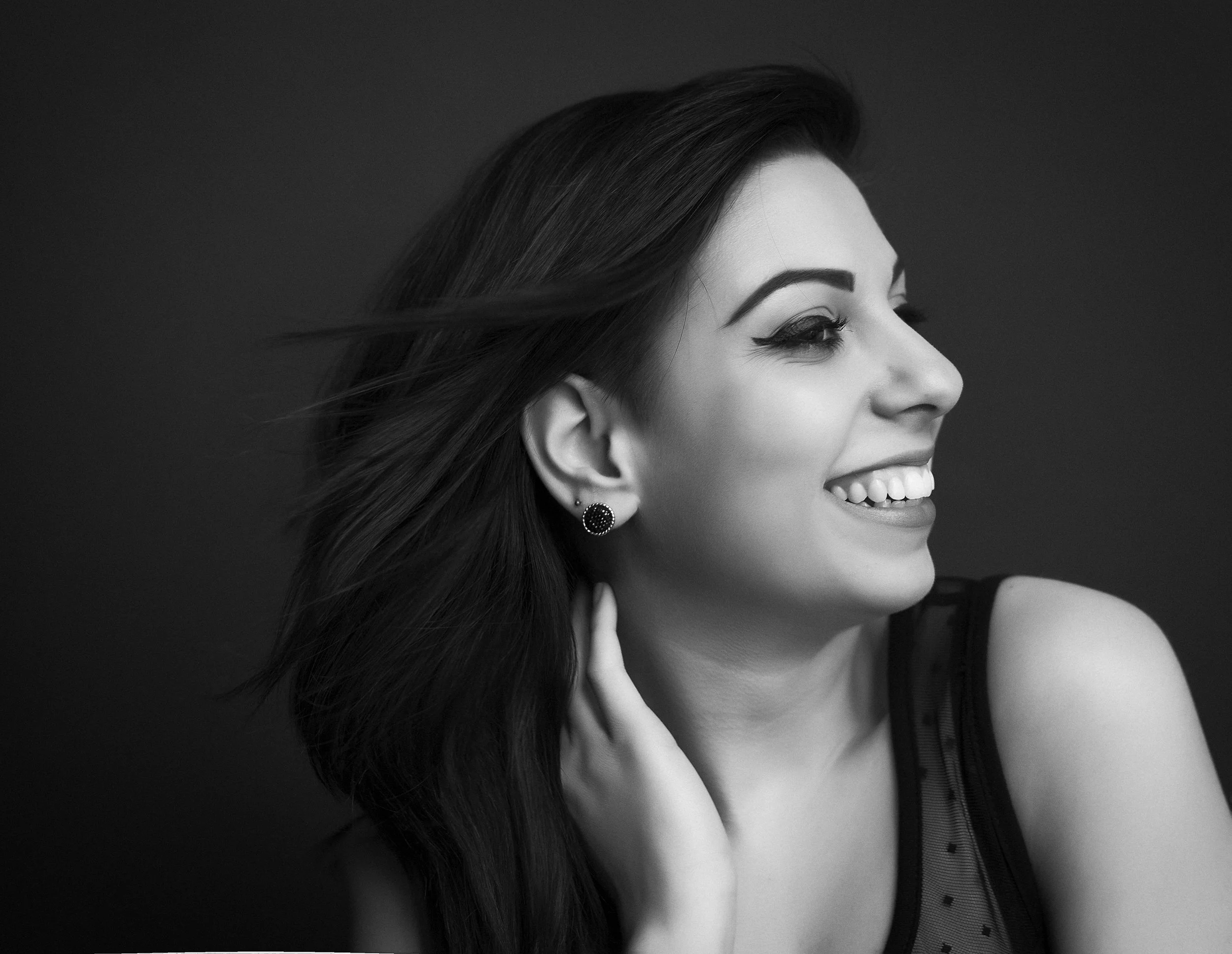 A black and white portrait of a smiling woman with long hair, wearing earrings and a sleeveless top.