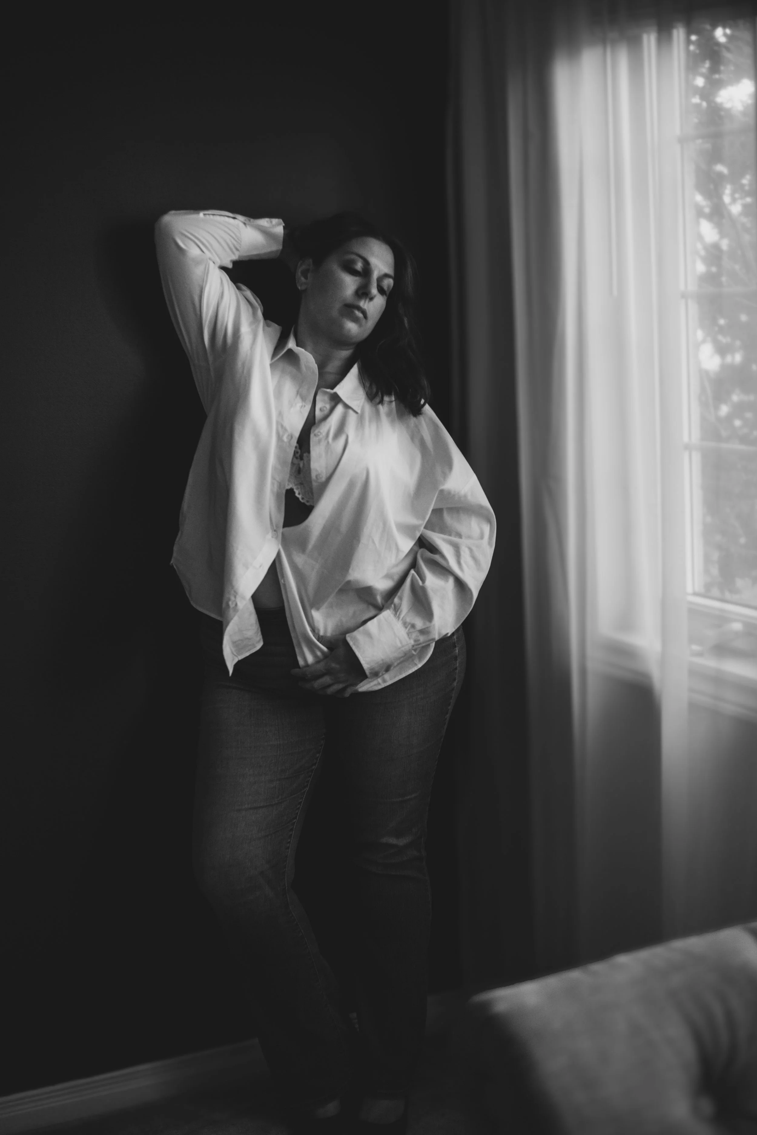 Black and white photo of a woman standing indoors near a window with light curtains, with her eyes closed and one arm raised behind her head, wearing a white shirt and jeans.