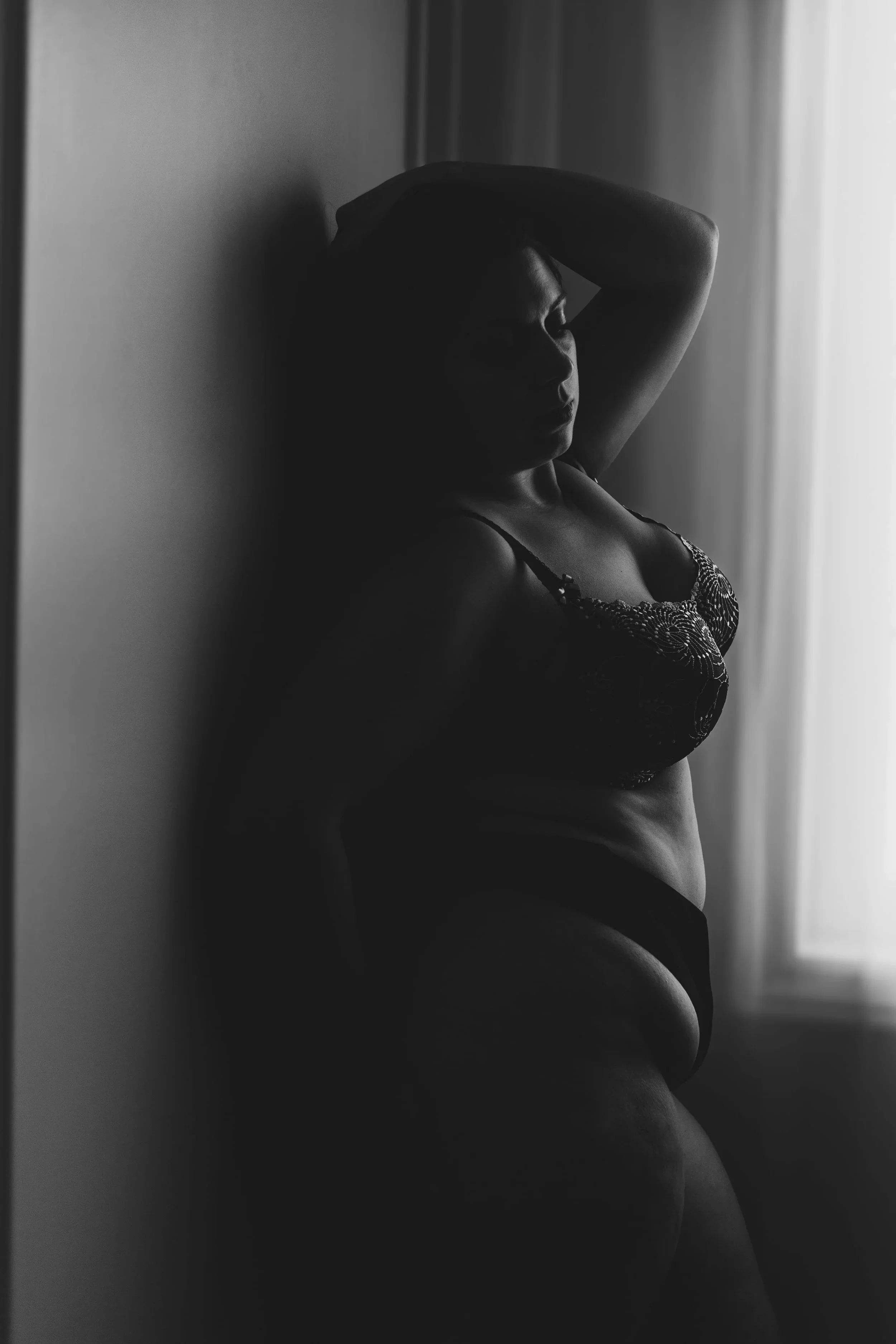 Black and white photo of a woman with her eyes closed, standing sideways near a window, with one arm resting on her head, wearing a lace bra and lingerie.