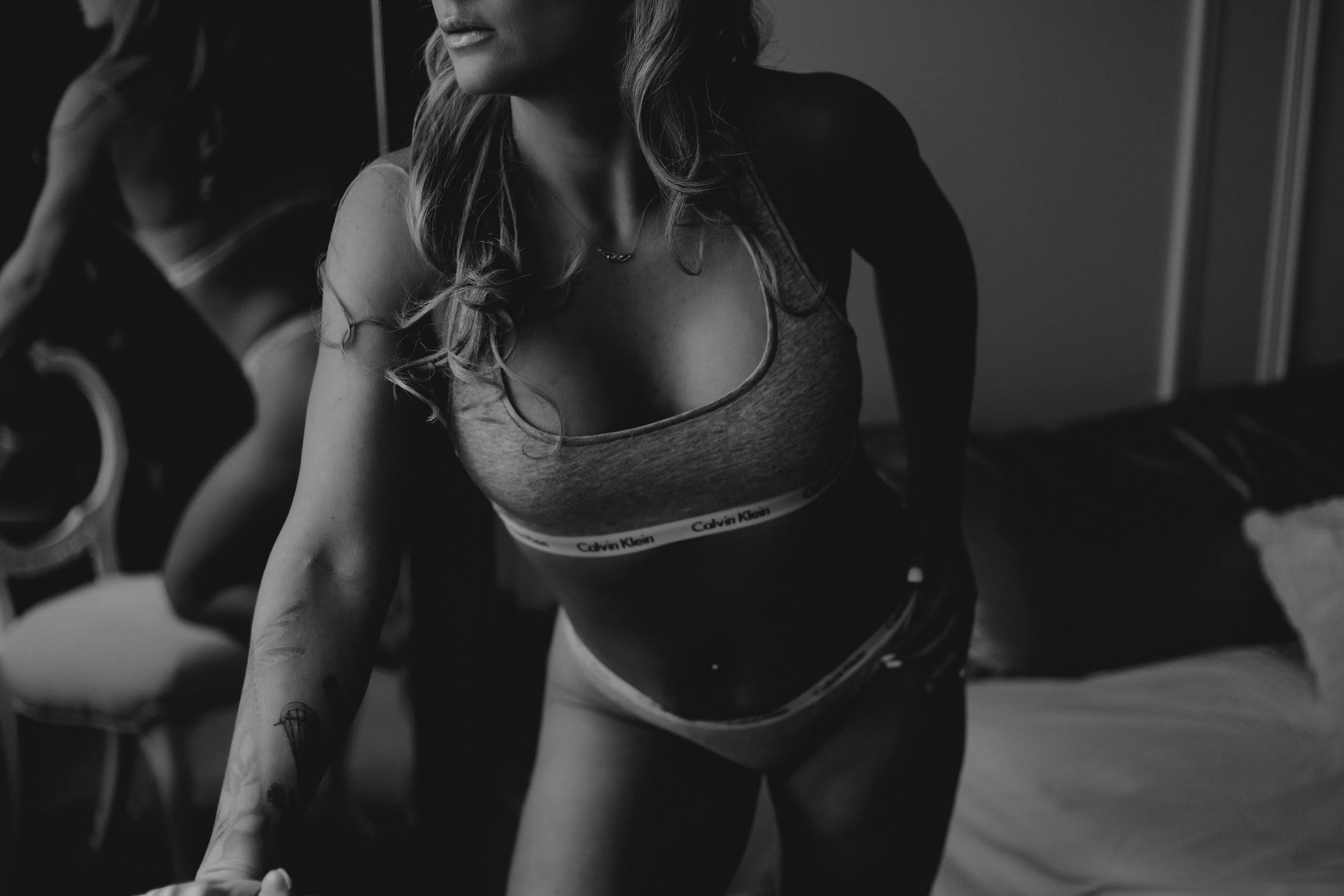 Black and white photo of a woman in a sports bra and shorts, leaning forward with her left arm extended, showing tattoos on her forearm. Her face is partially visible, with her hair flowing over her shoulders. She appears to be in a room with a guita