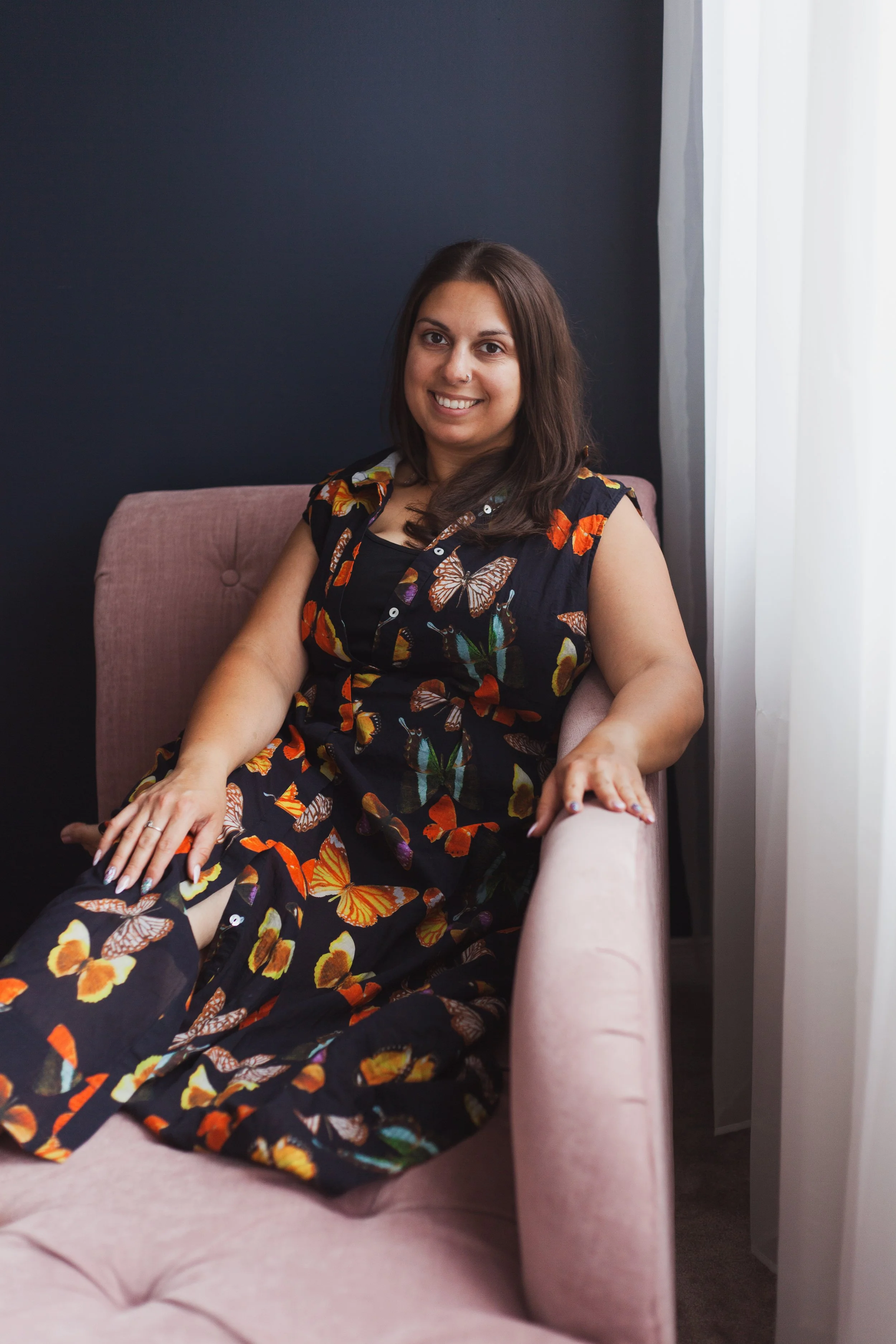A woman with dark hair, sitting on a pink chair in front of a dark wall and a white curtain, wearing a black dress with colorful butterfly patterns, smiling at the camera.