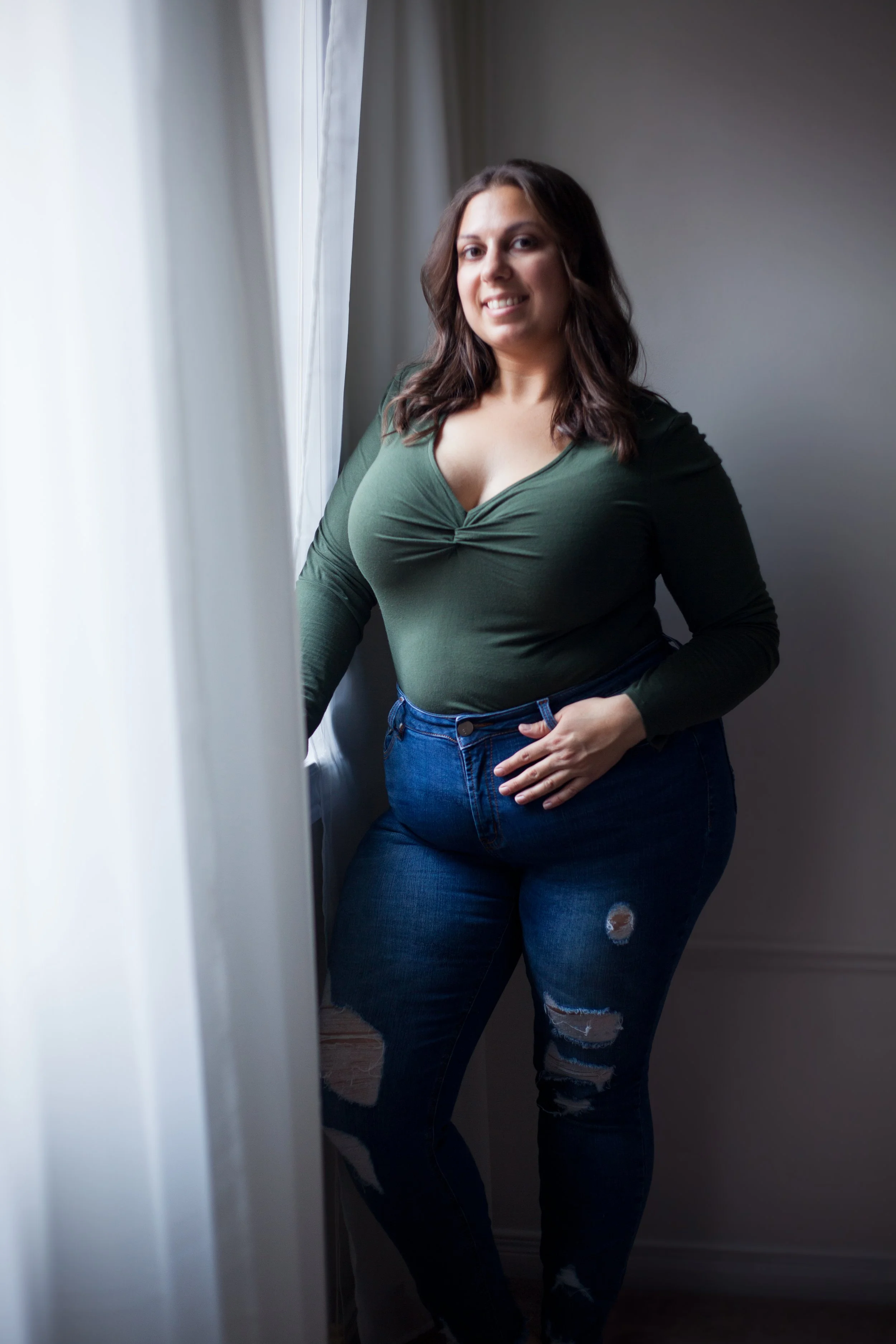 A woman with shoulder-length brown hair wearing a dark green long-sleeved shirt and ripped jeans standing near a window with curtains indoors.