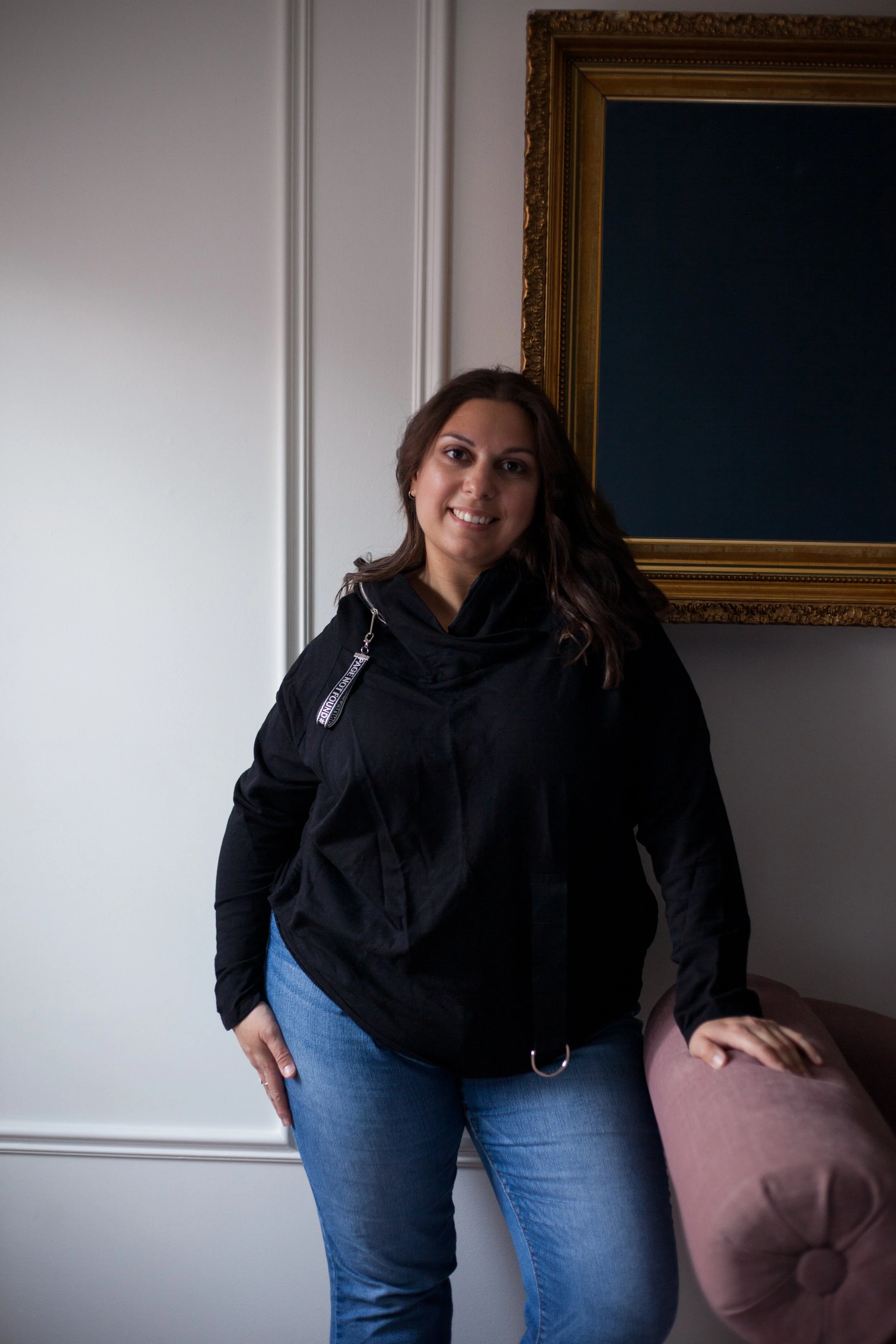 A woman with long wavy brown hair wearing a black jacket and blue jeans, smiling and standing in front of a white wall with decorative molding and a dark ornate picture frame.