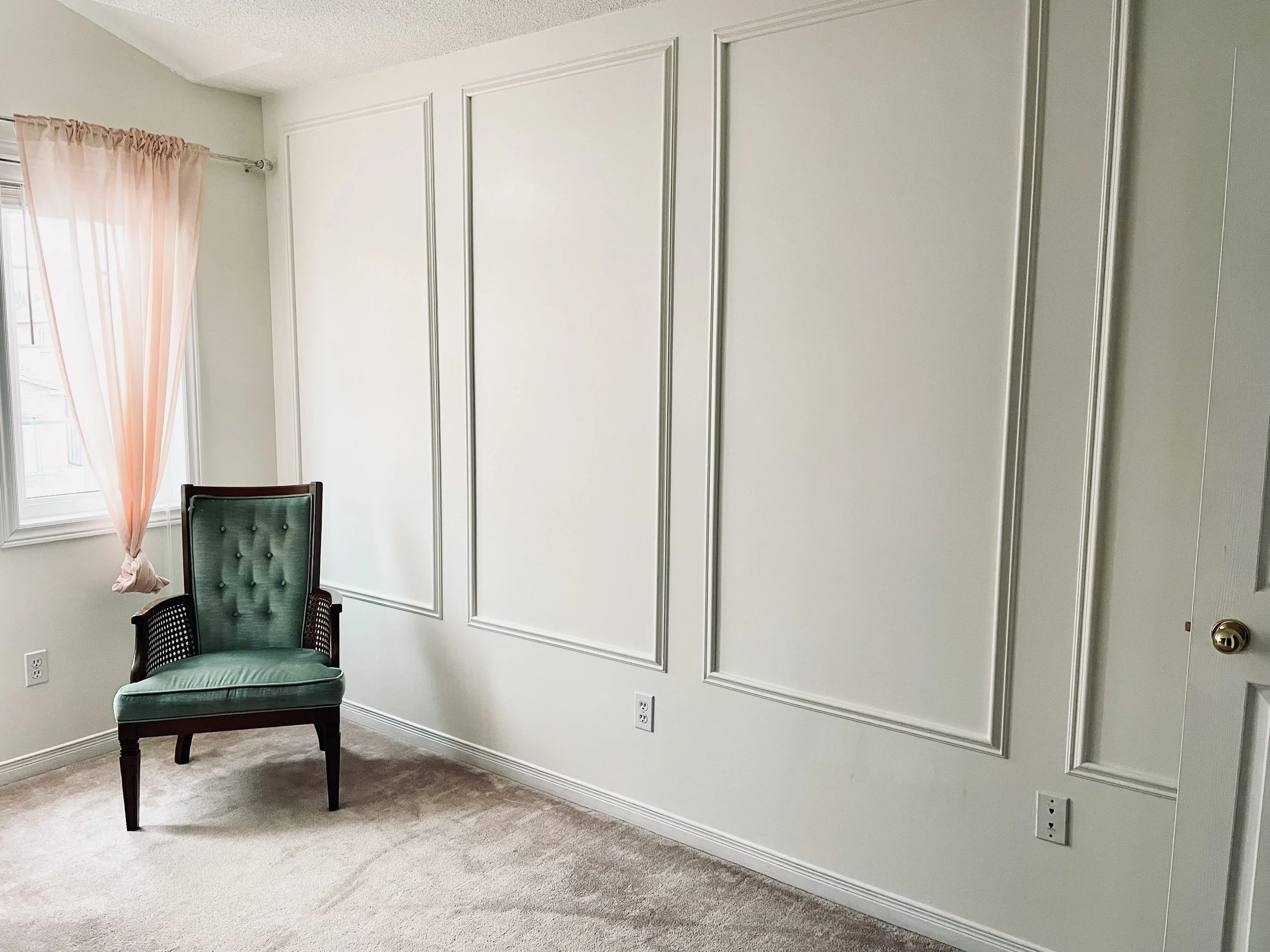 A room with a window covered by light pink curtains, a green upholstered chair with dark wood arms, beige carpet, and a white wall with decorative molding and outlets.