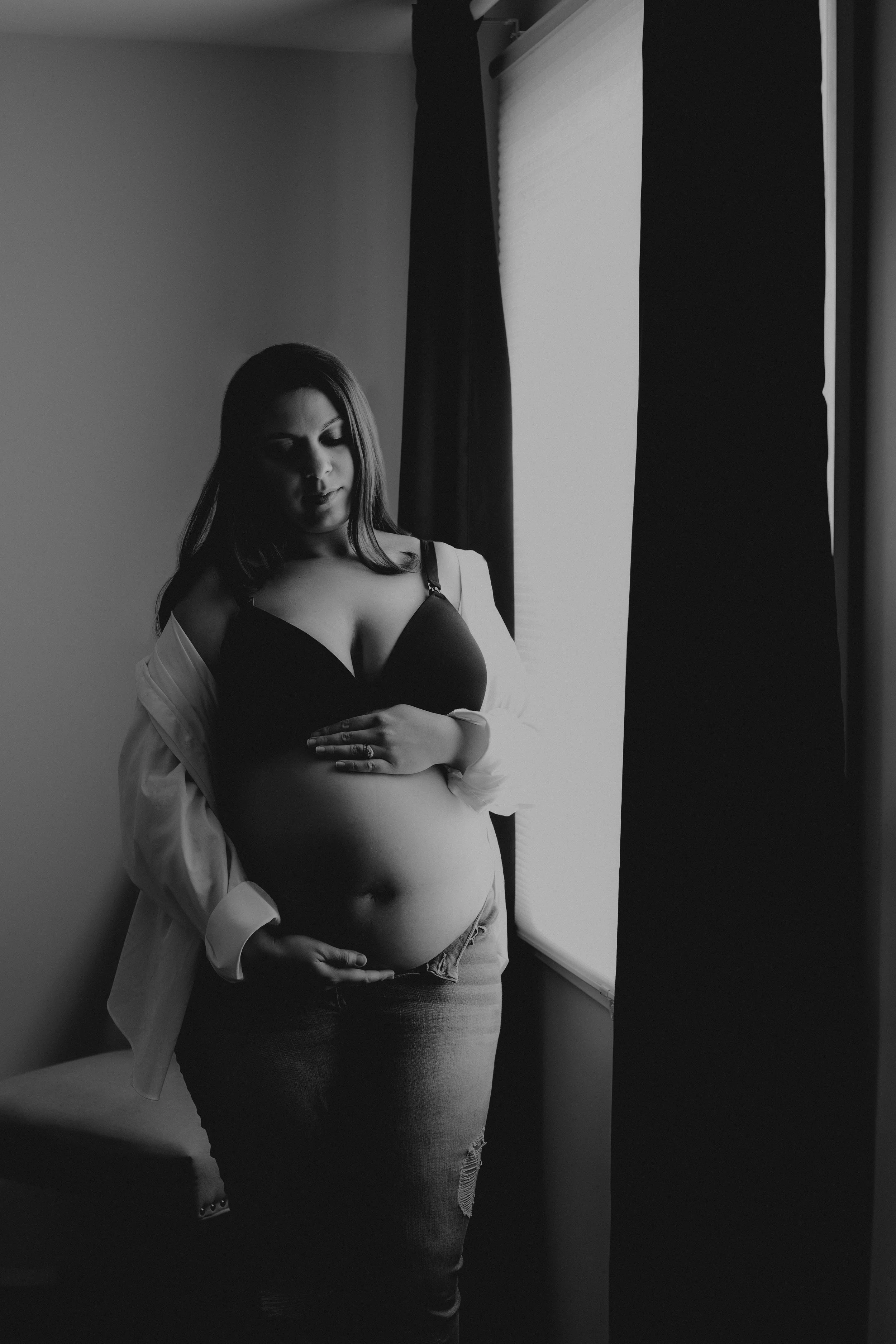 A woman with long hair wearing a black bra, jeans, and a white shirt draped off her shoulders, standing near a window with curtains. She is cradling her pregnant belly with one hand and her eyes are closed.