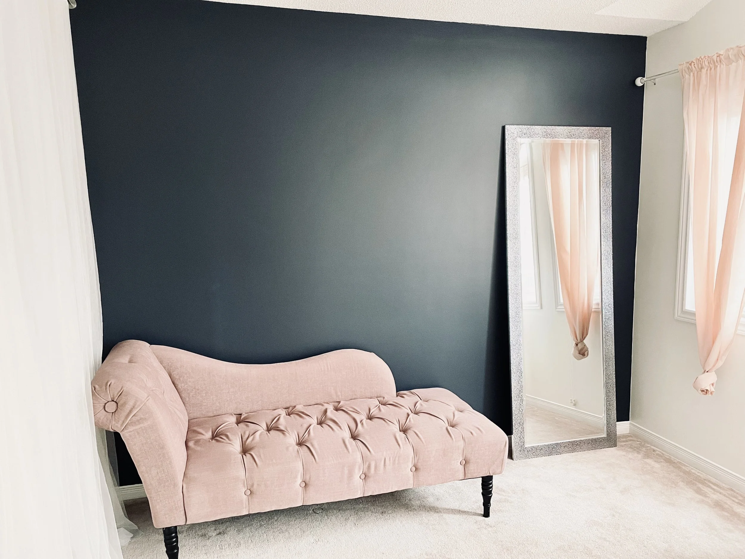 A pink upholstered tufted bench with black turned legs in front of a large silver-framed mirror, next to a window with light pink curtains in a room with a dark accent wall.
