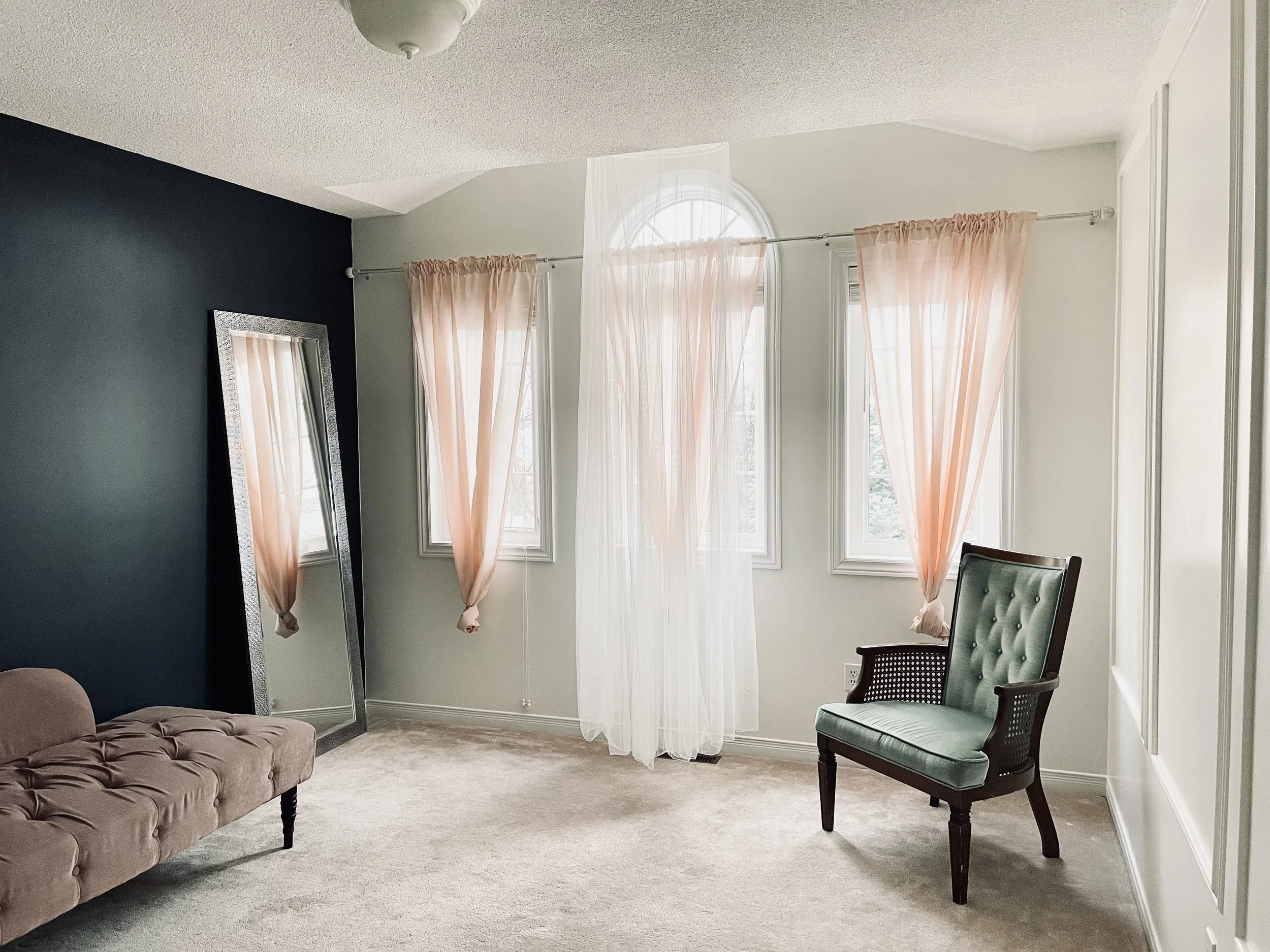 Living room interior with beige tufted sofa, green upholstered chair, large mirror, and windows with pink and sheer curtains.