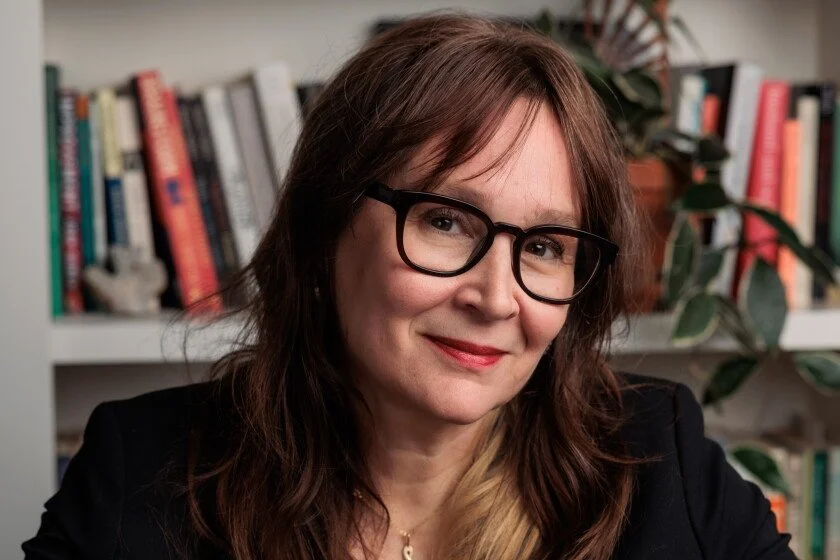 Writer and editor, Susannah Felts. Headshot features her wwith shoulder-length brown hair, wearing black glasses and a black blazer, smiling in front of a bookshelf with colorful books and a potted plant.