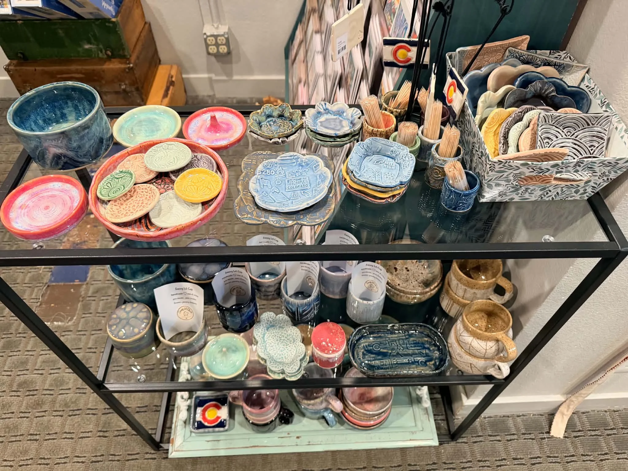 Display table with handmade pottery and ceramic dishes, bowls, cups, and various craft items in a retail shop.