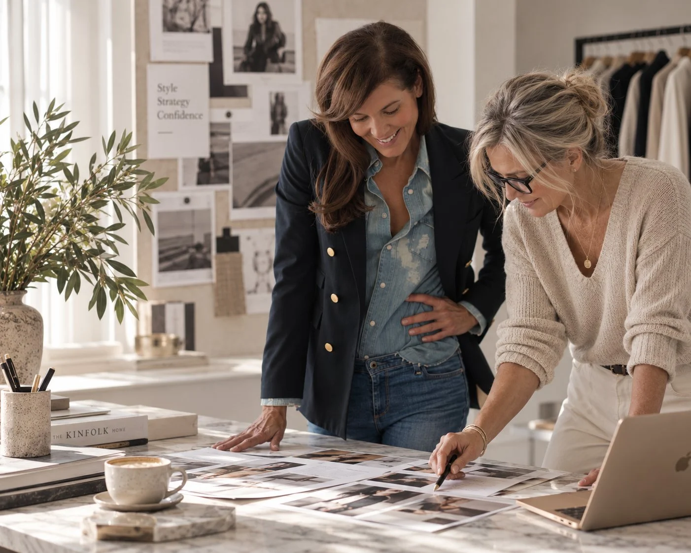 Two women working together at a marble table, reviewing photos and design plans, surrounded by a plant and artsy decor. One woman is pointing at a photo on the table.