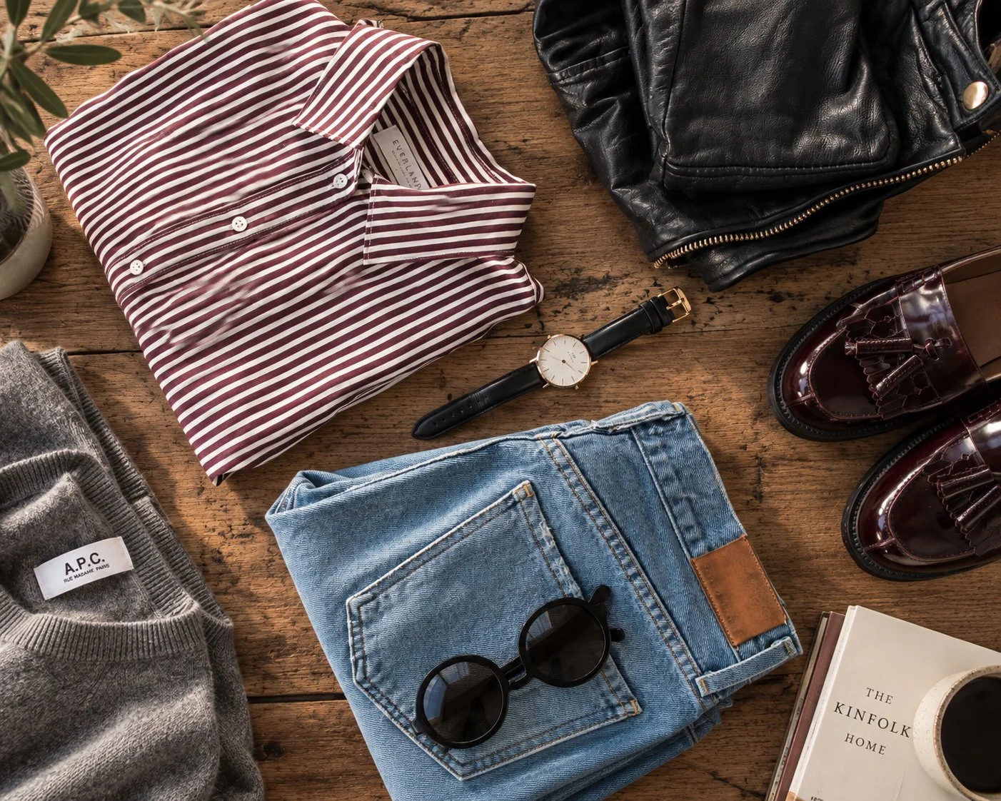 Flat lay of men's fashion items on a wooden surface, including a folded red and white striped shirt, a pair of blue jeans with black sunglasses on top, a black watch, a black leather jacket, burgundy loafers, a gray sweater, a book titled 'The Kinfolk Home,' and a white mug.