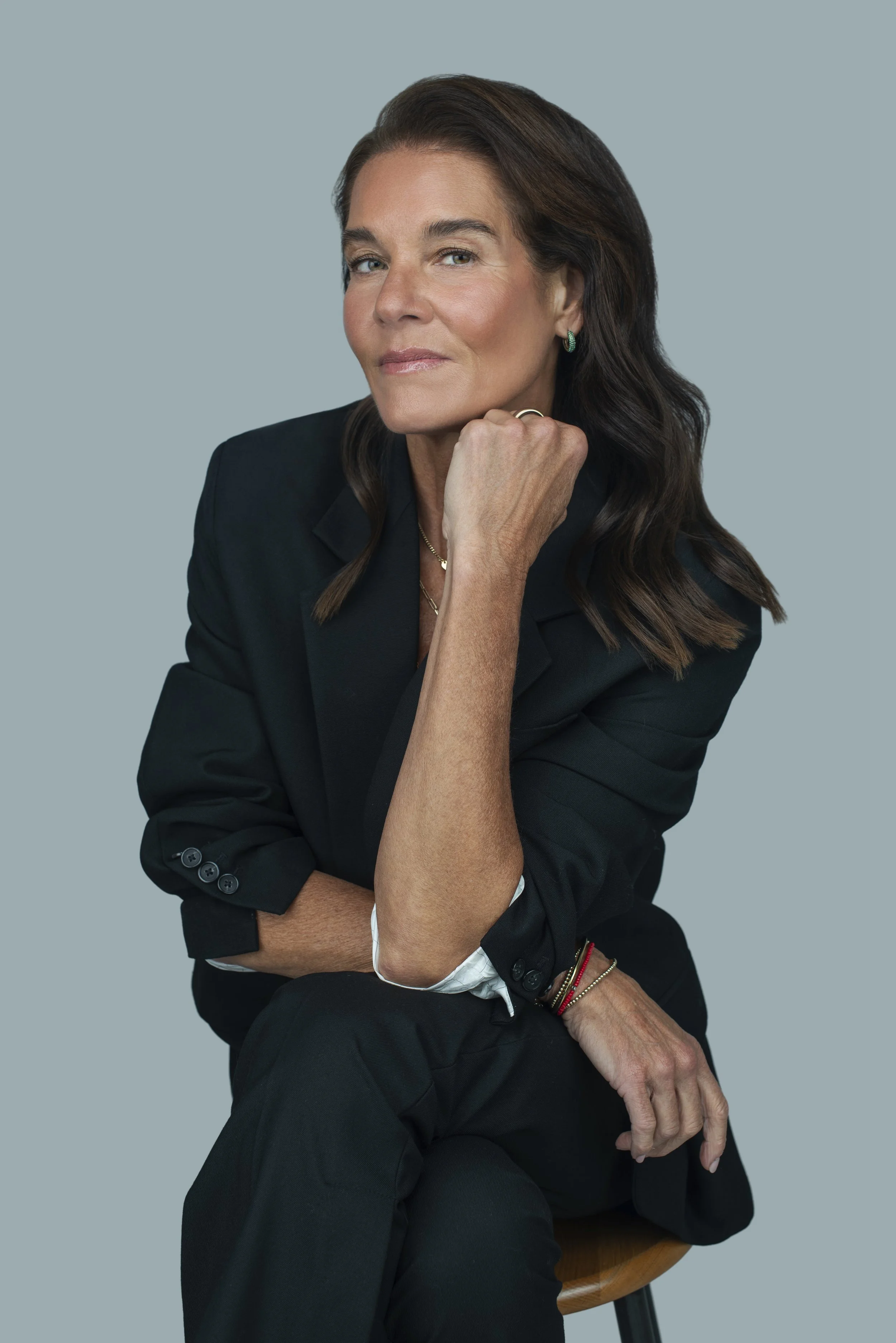 A confident middle-aged woman with long brown hair, wearing a black blazer, jewelry, and makeup, sitting on a wooden stool against a grey background.