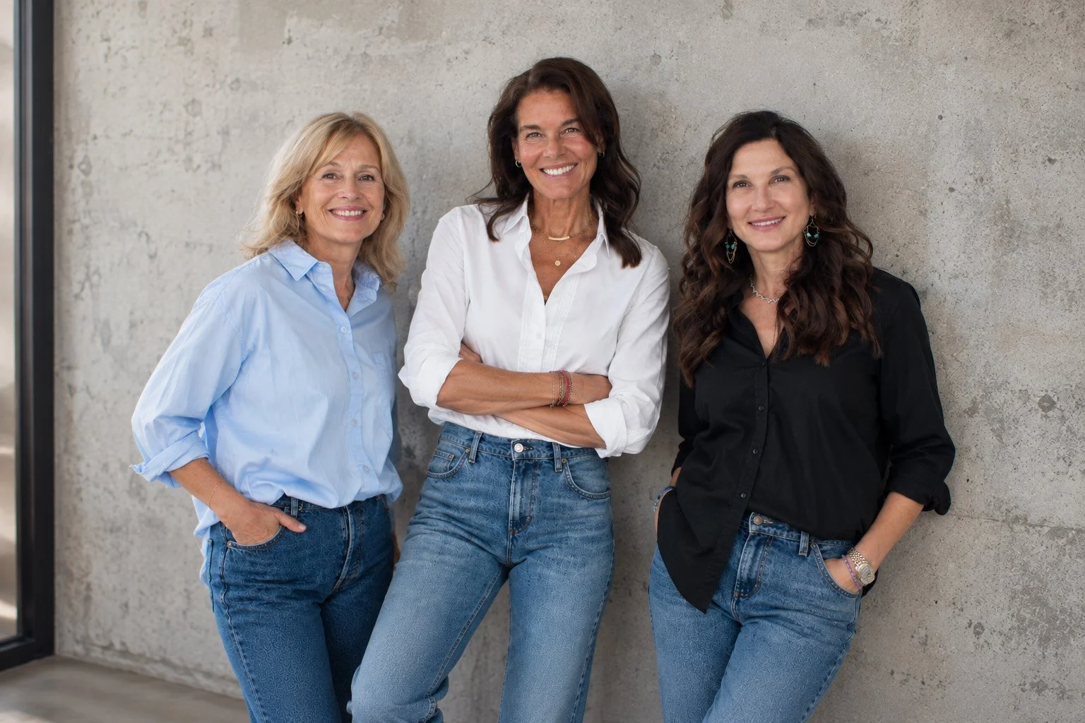 Three women standing together against a concrete wall, smiling, dressed in casual shirts and jeans.