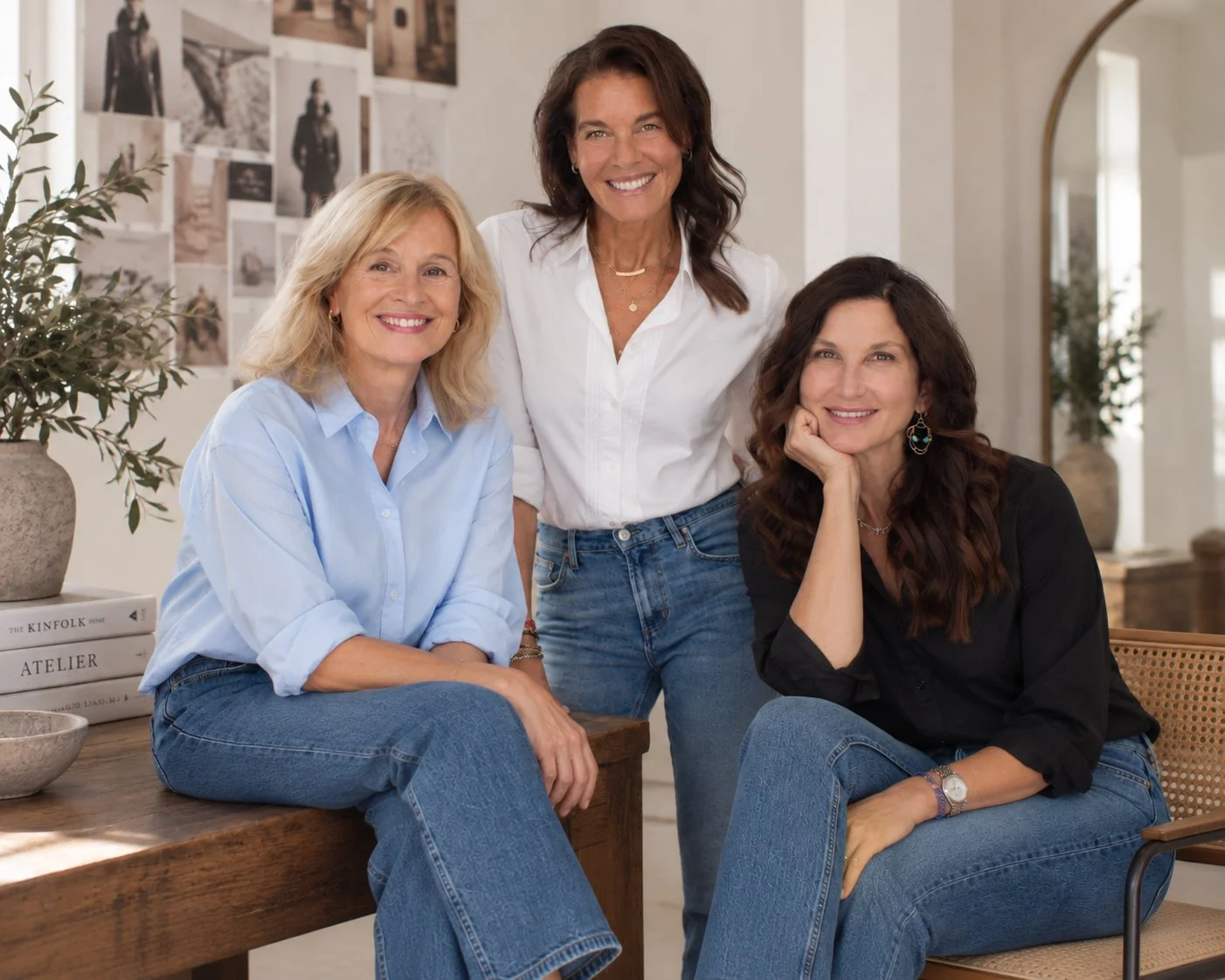 Four women smiling in a bright, cozy room, some sitting and one standing behind, with photo collage on the wall and plants nearby.