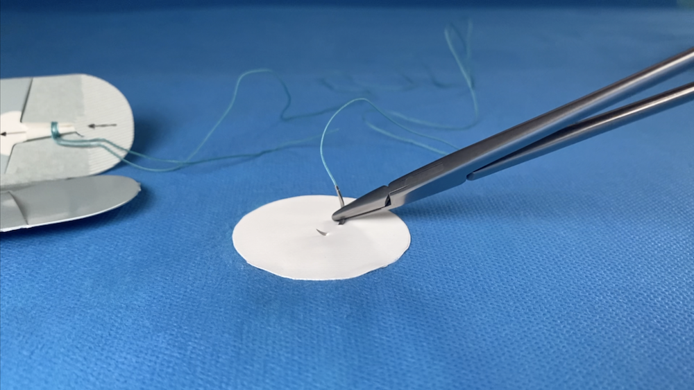 Close-up of surgical tools, including forceps holding a fine thread, on a blue surgical drape, with a medical device in the background.