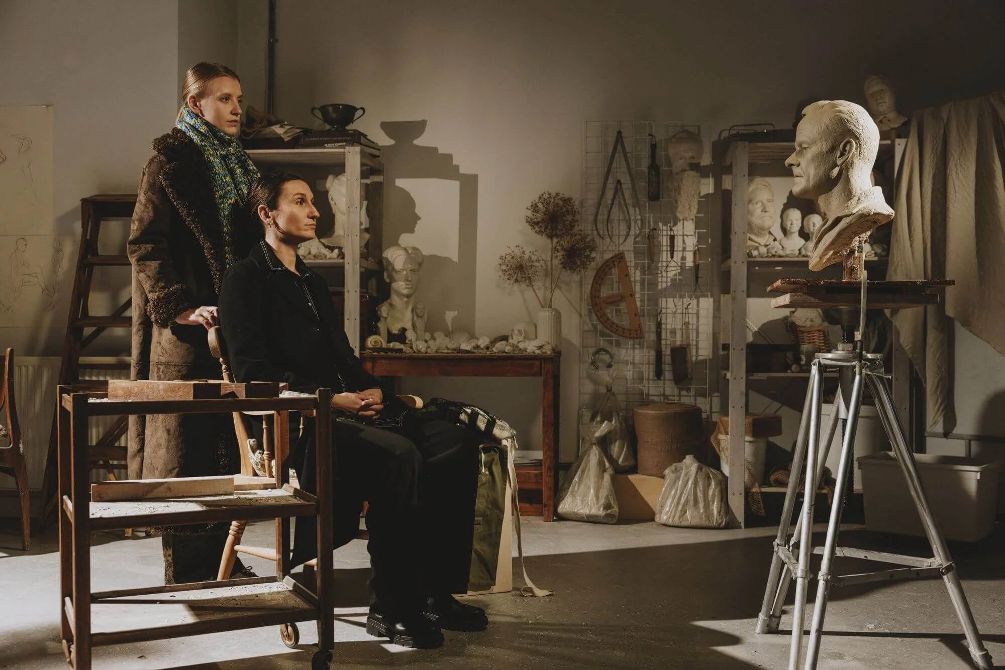 Bessie Carter and Poppy Lee Friar in a sculpture studio surrounded by busts, tools and works in progress.