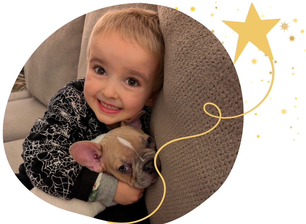 A young boy with blonde hair, wearing a black shirt with white spiderweb pattern, smiling while hugging a small beige French Bulldog puppy with a white marking on its forehead, in a cozy living room.