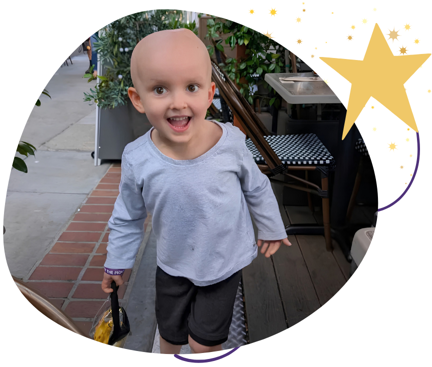 A young child with a bald head smiling and looking at the camera, standing outdoors near a brick pathway and surrounded by plants.