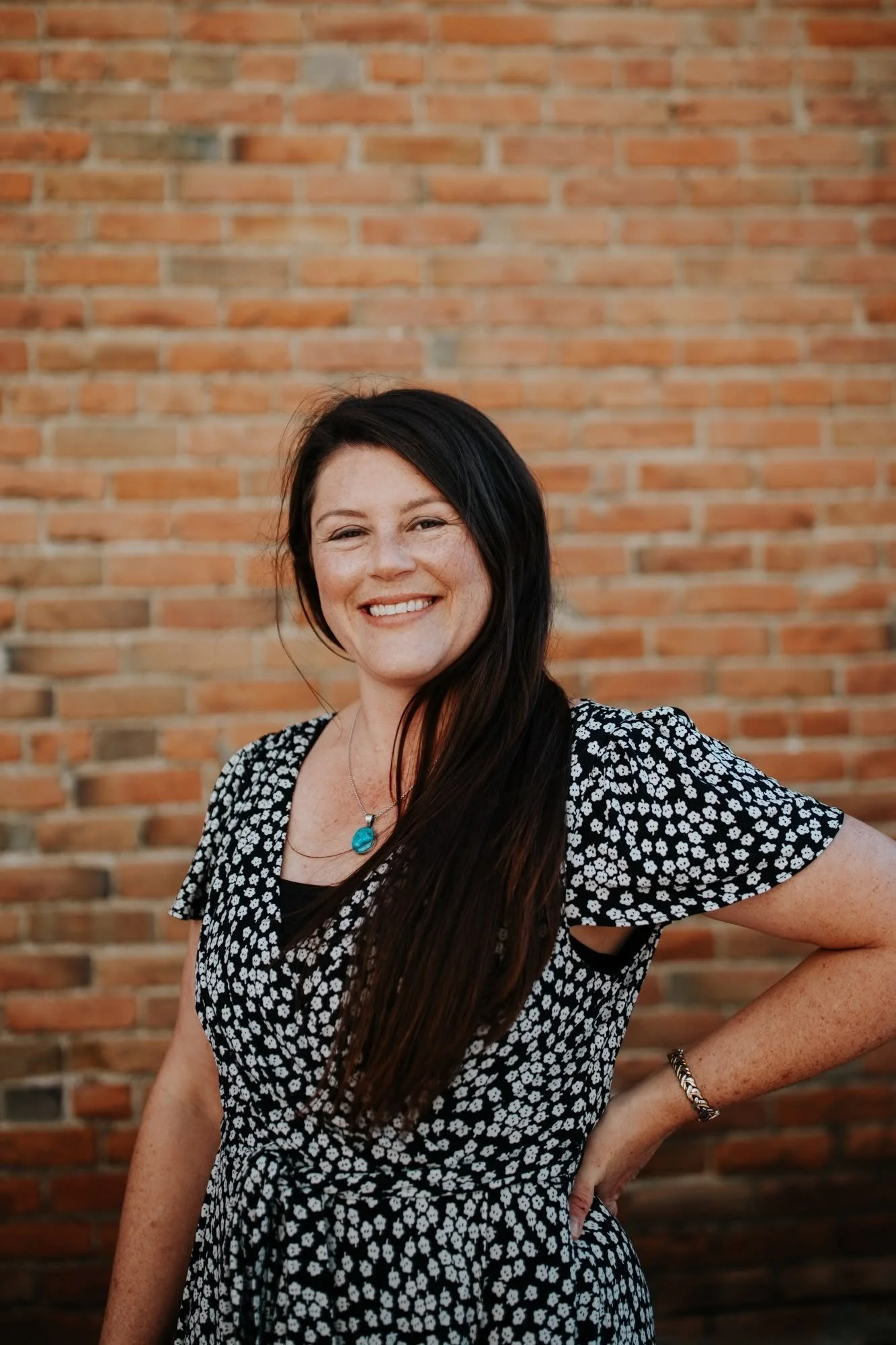 A woman with long dark hair smiling in front of a brick wall, wearing a black and white floral dress, a turquoise necklace, and a bracelet.