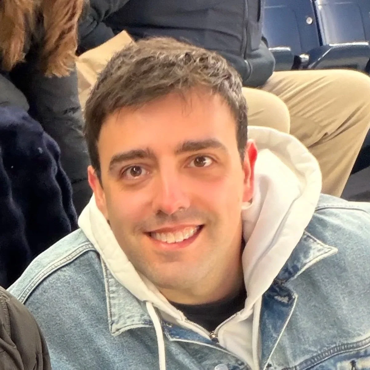 A smiling man with short dark hair wearing a white hoodie under a denim jacket, sitting in a crowded indoor space.