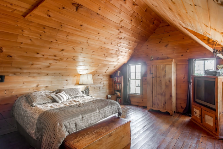 Cozy bedroom with wood-paneled walls and ceiling, a bed with patterned bedding, a wooden chest at the foot of the bed, a small bookshelf, a window with curtains, a wardrobe, a lamp on bedside table, and a wooden TV stand with an old-style TV.