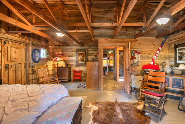 Interior of a rustic log cabin bedroom with wooden walls and ceiling, a bed with a quilt, a rocking chair, a dresser with a lamp, and a doorway leading to another room. Decorations include a mirror, a picture, and a flag with a red, white, and blue theme.