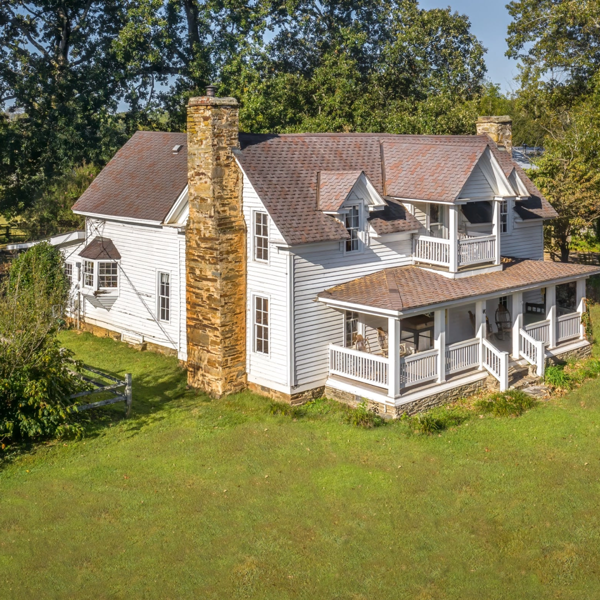 A large two-story white house with a brick chimney, multiple gabled roofs, a front porch with white railing, and surrounded by greenery.