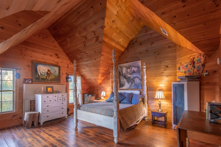 A cozy bedroom with wooden walls and ceiling, featuring a four-poster bed with a bedspread, two nightstands with lamps, a dresser, a painting of a horse, and a window showing greenery outside.