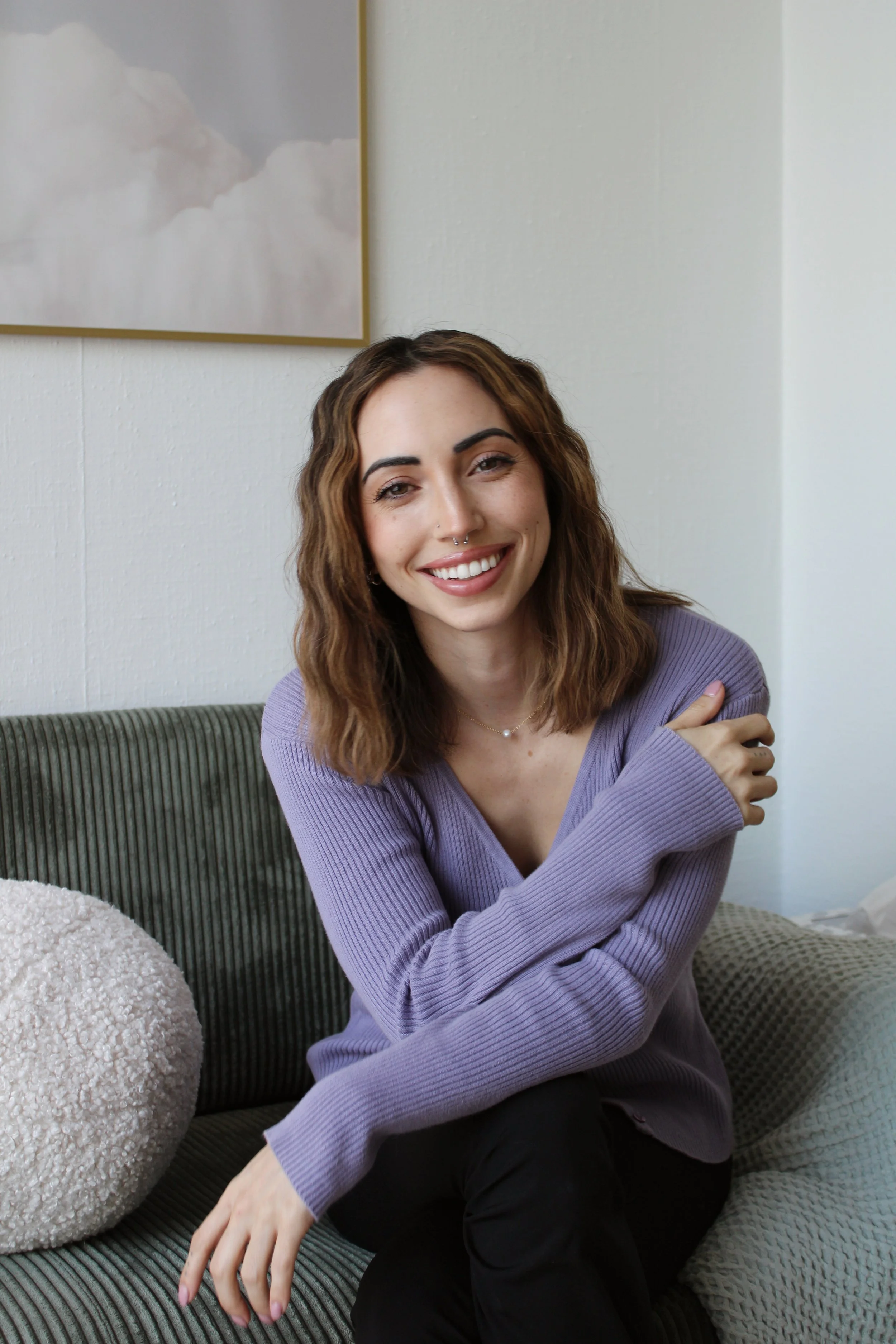 A woman with wavy brown hair, wearing a purple ribbed sweater, smiling and sitting on a green textured couch, with a cream fuzzy pillow and a gray pillow nearby, against a white wall with a framed picture of clouds.
