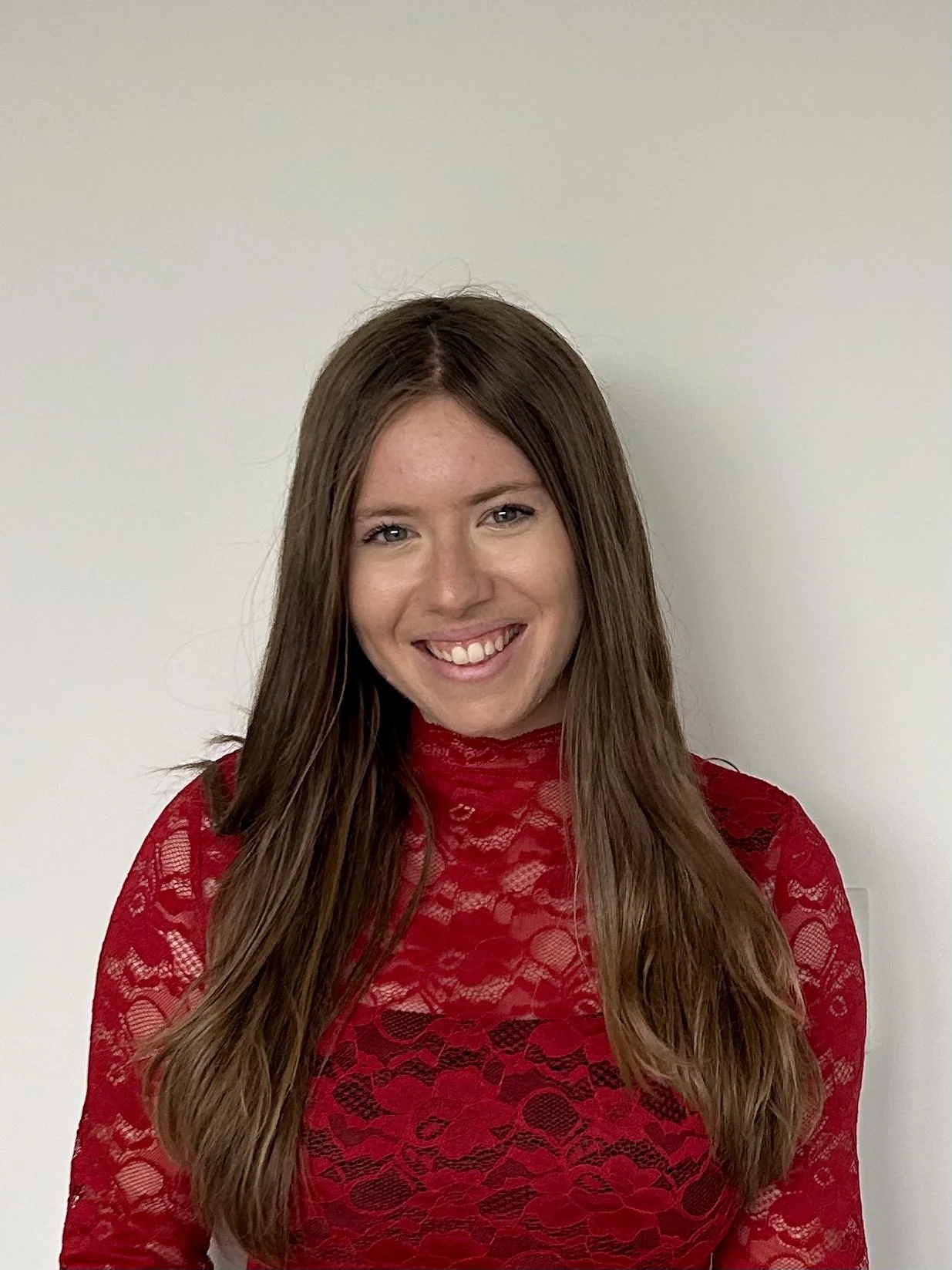 A woman with long brown hair smiling, wearing a red lace top, standing against a plain white wall.
