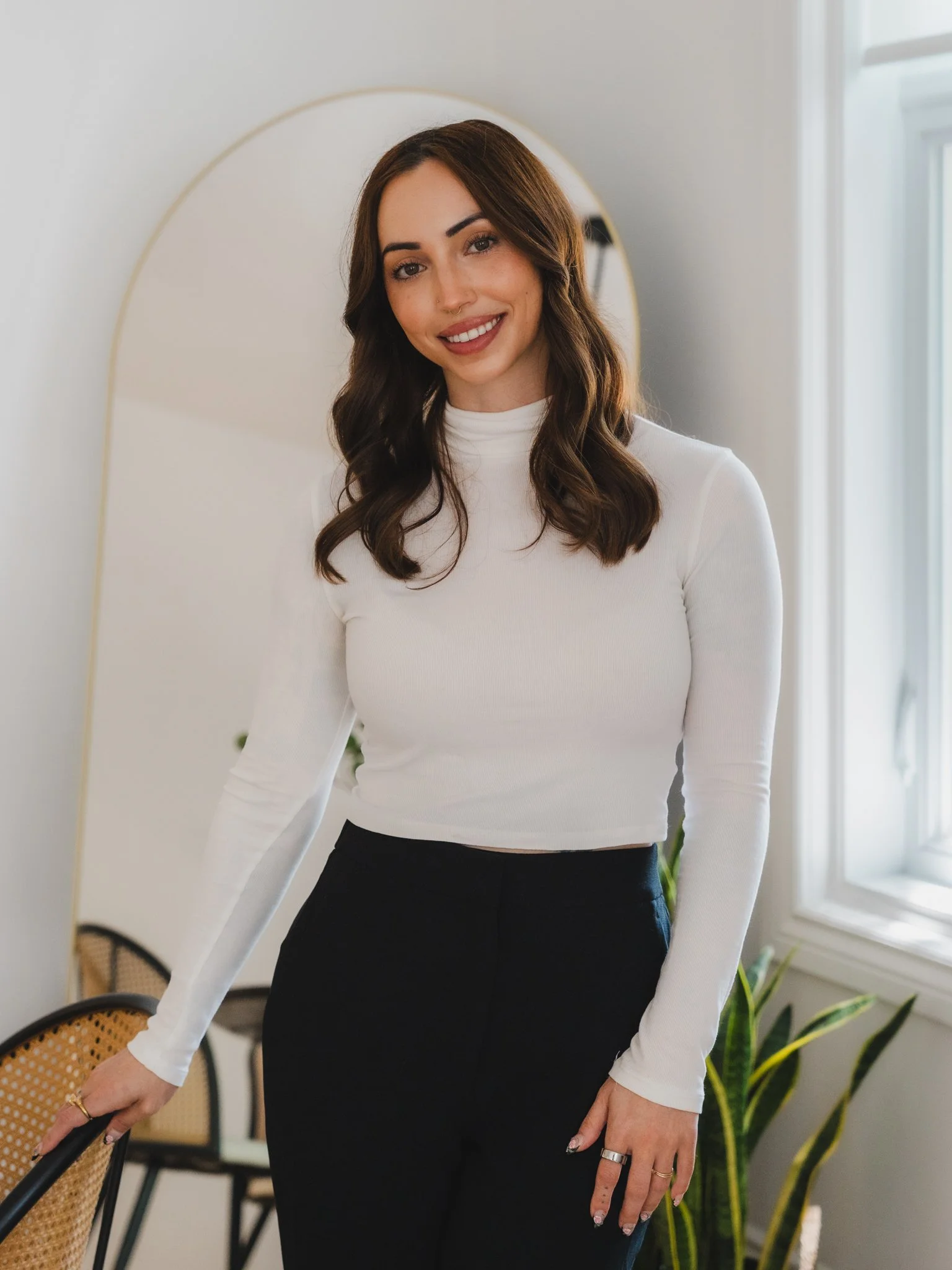 A woman with brown hair, wearing a white long-sleeve top and black pants, standing beside a chair with a cane backrest. She is smiling, with a neutral-colored wall and window in the background.