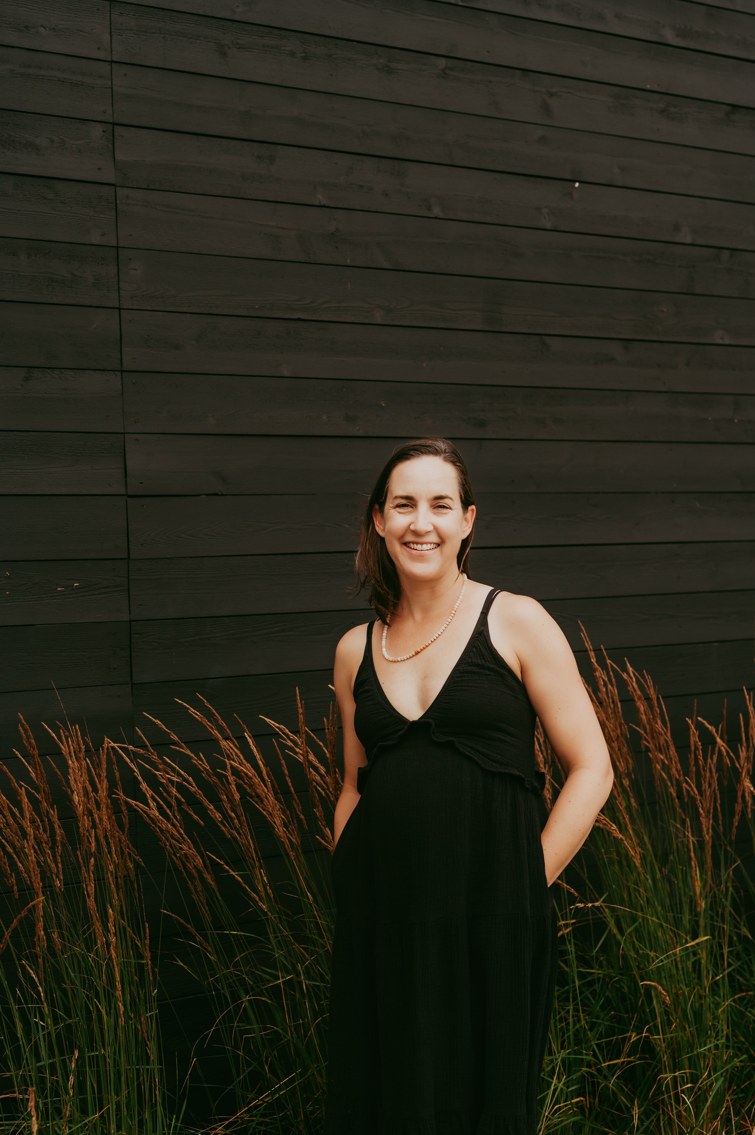 A woman with shoulder-length brown hair smiling and wearing a black dress with spaghetti straps, standing outdoors in front of a dark wooden wall, surrounded by tall brown grasses.