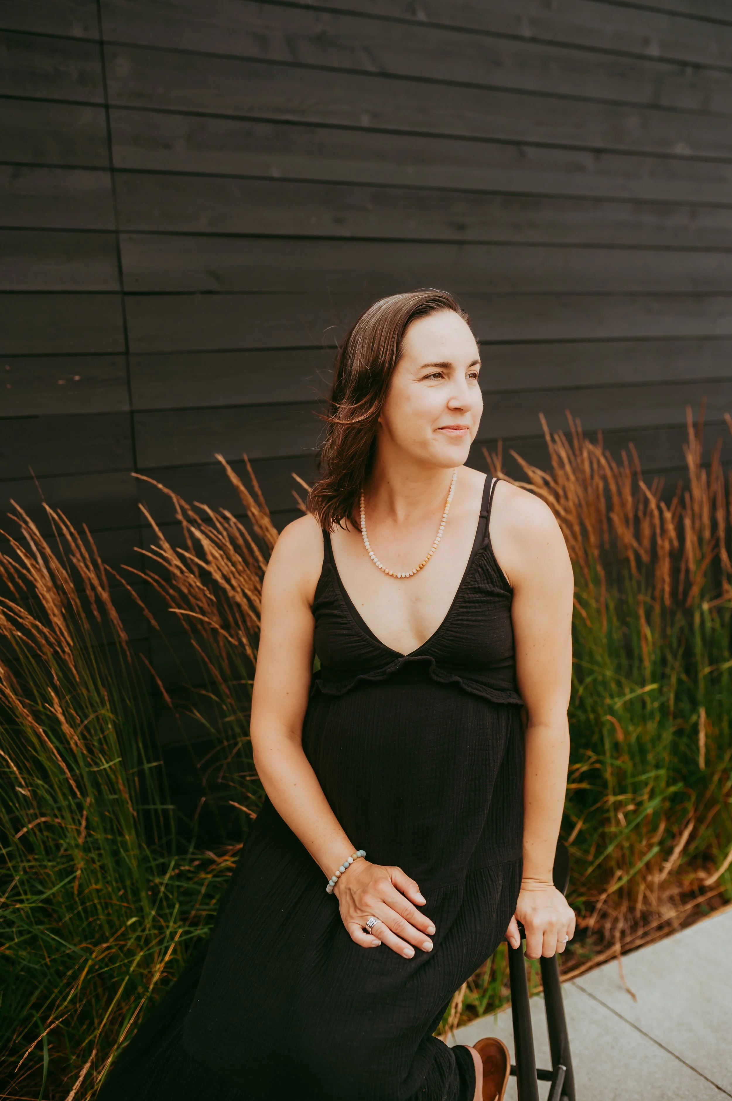 A woman sitting outside holds a cane and is surrounded by tall grasses, with a black wooden wall behind her.