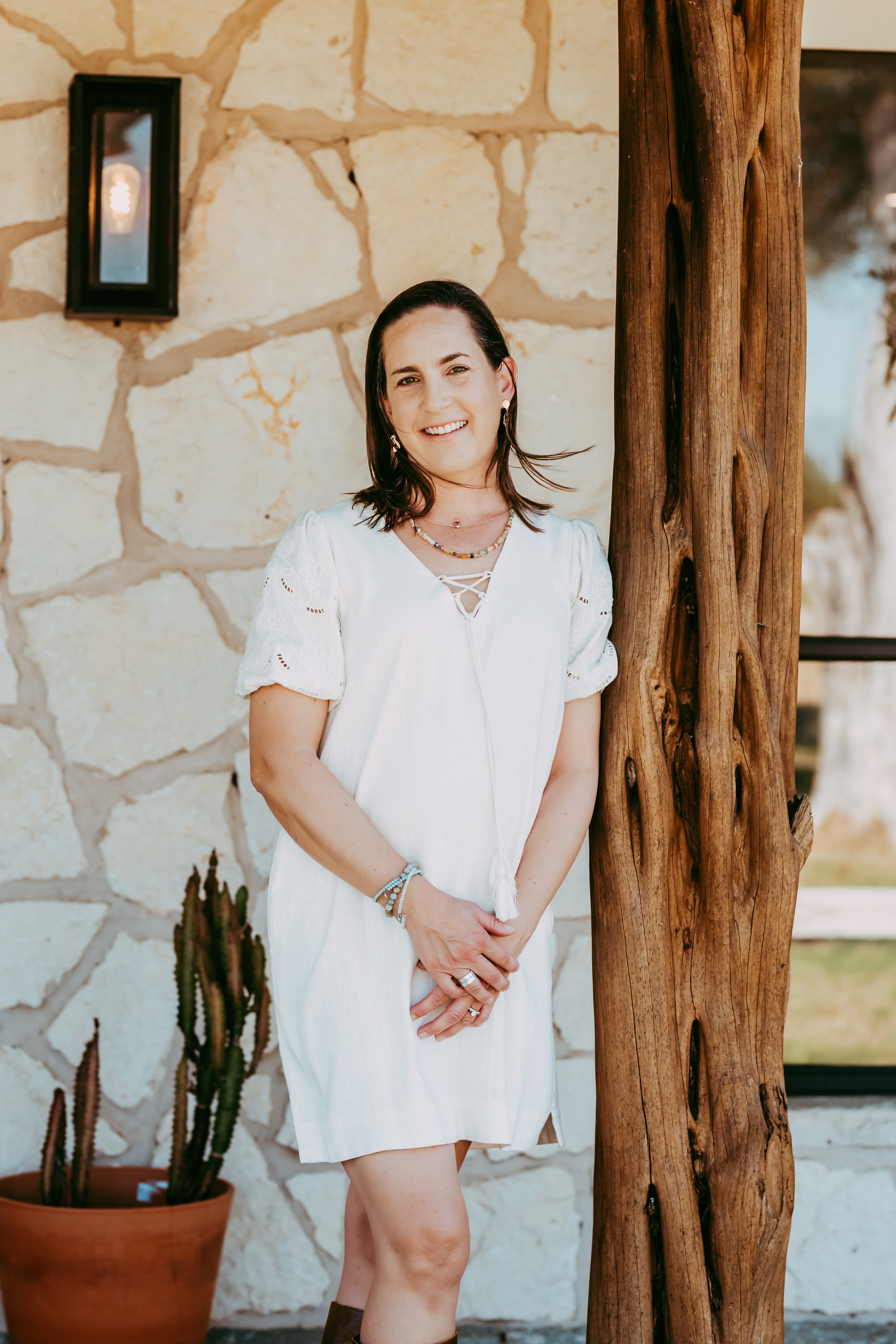 A woman with shoulder-length dark hair smiling while standing outdoors near a rustic wooden post, wearing a white dress with lace details, jewelry, and a bracelet. Behind her is a stone wall and a potted cactus plant.