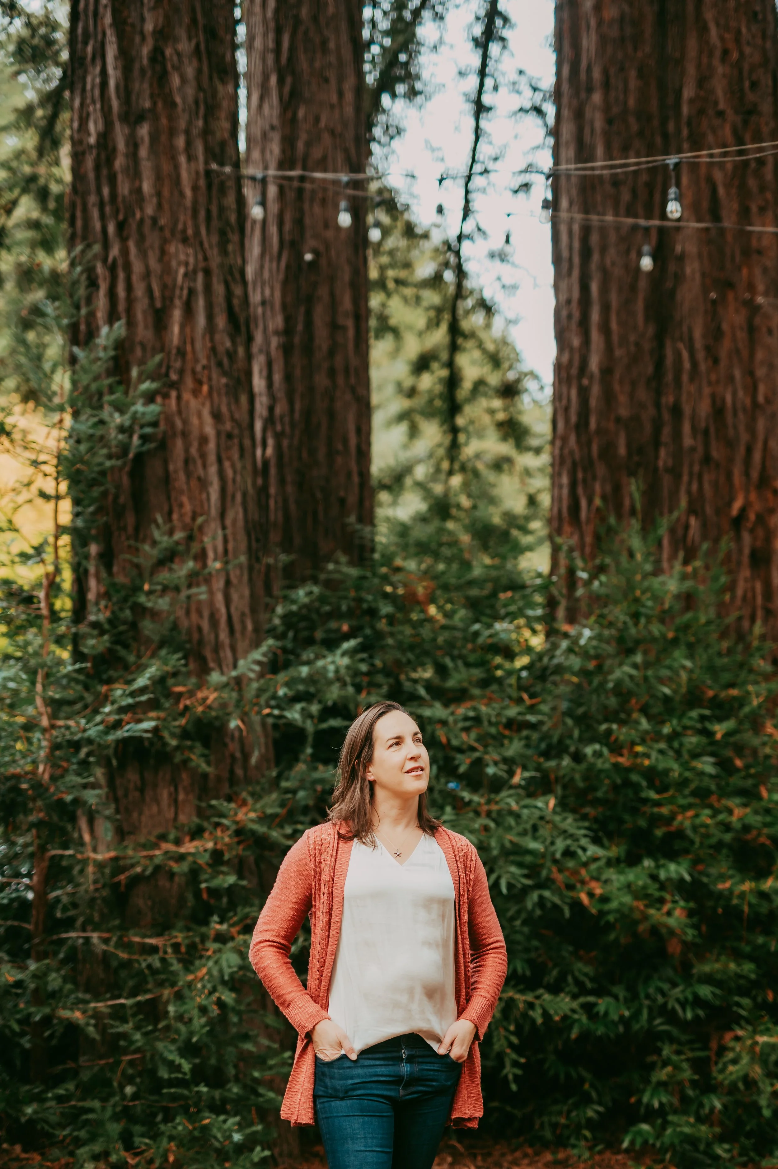 A woman with shoulder-length brown hair wearing a white shirt, an orange sweater, and blue jeans, standing outdoors in front of large redwood trees and greenery, looking upward.
