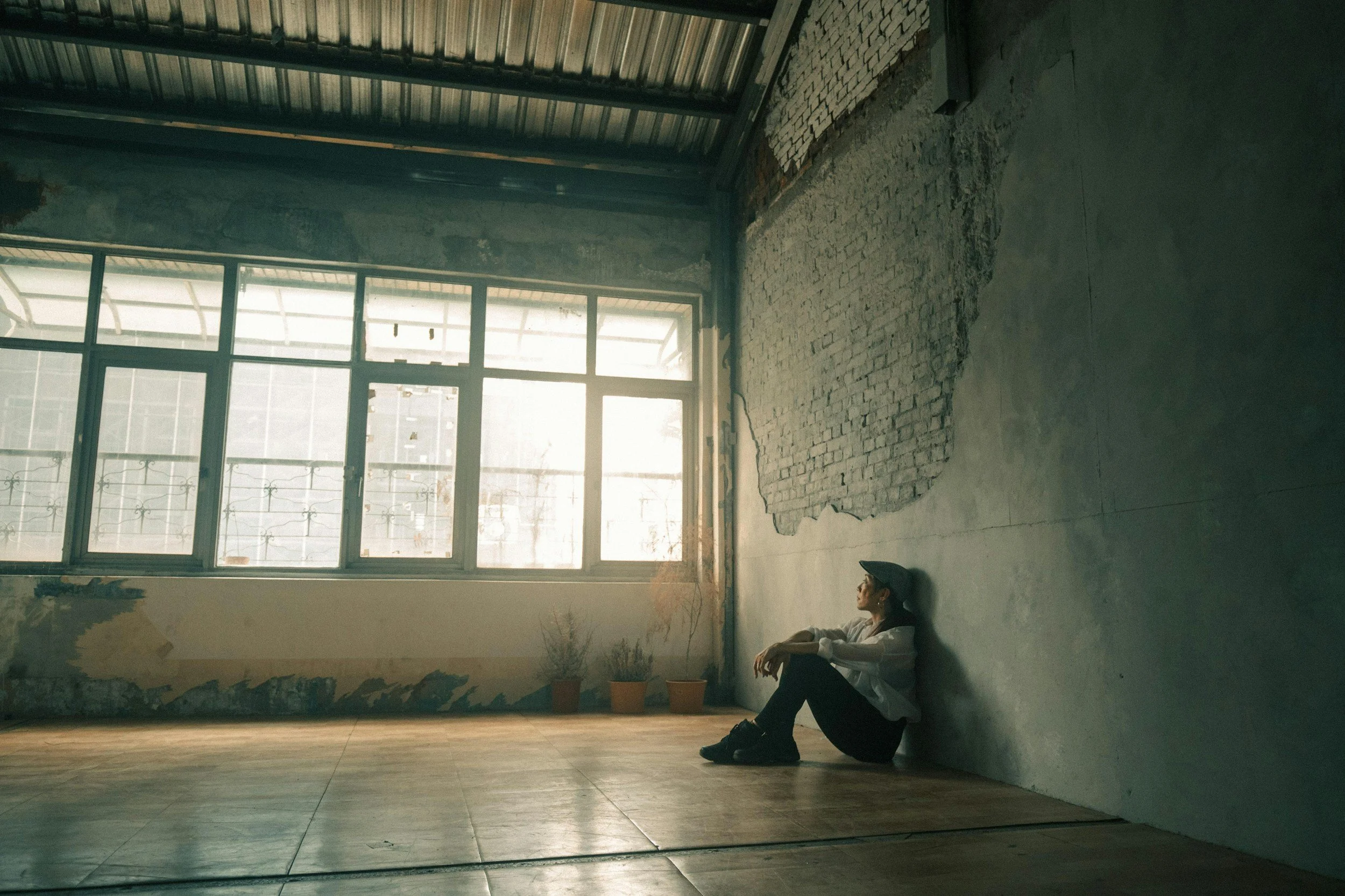 A woman sitting on the floor inside an unfinished room with exposed brick wall and large window, looking towards the windows with sunlight streaming in.