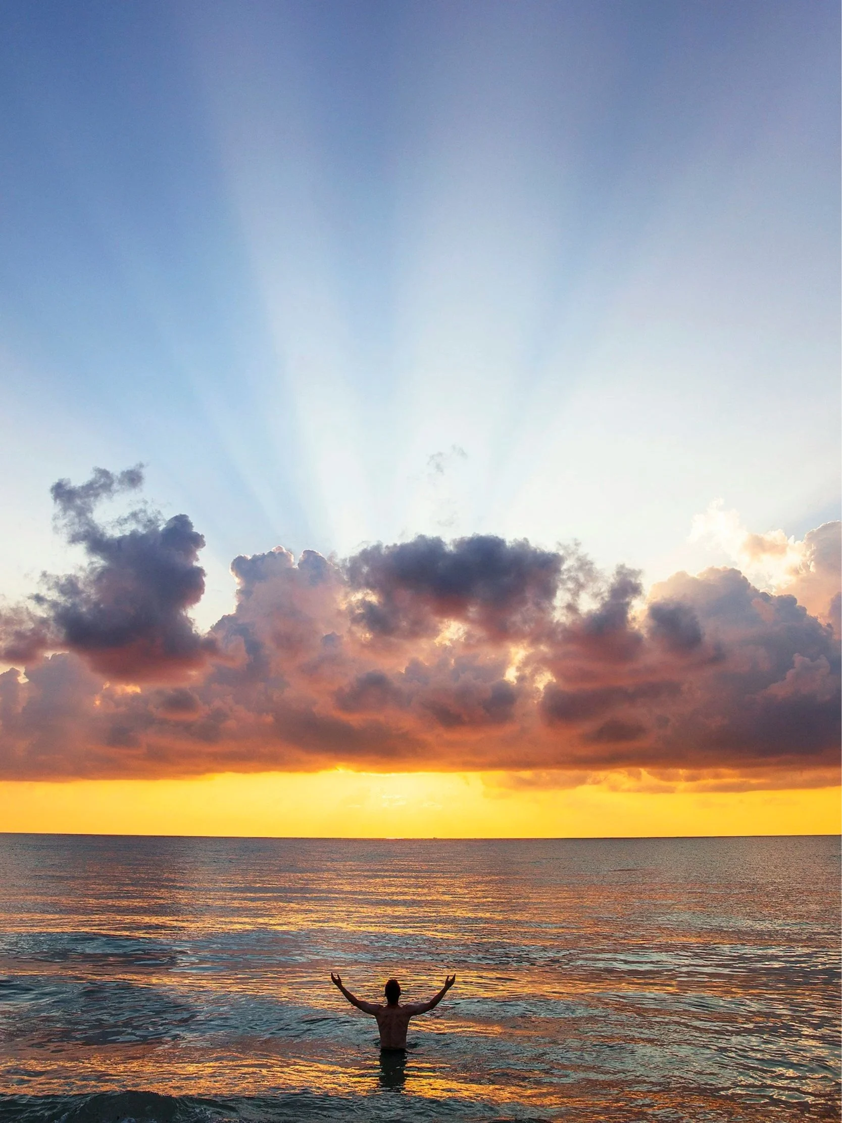 Person standing in ocean water with arms raised during a colorful sunset, reflecting orange and purple hues, with dark clouds above and a bright sky.