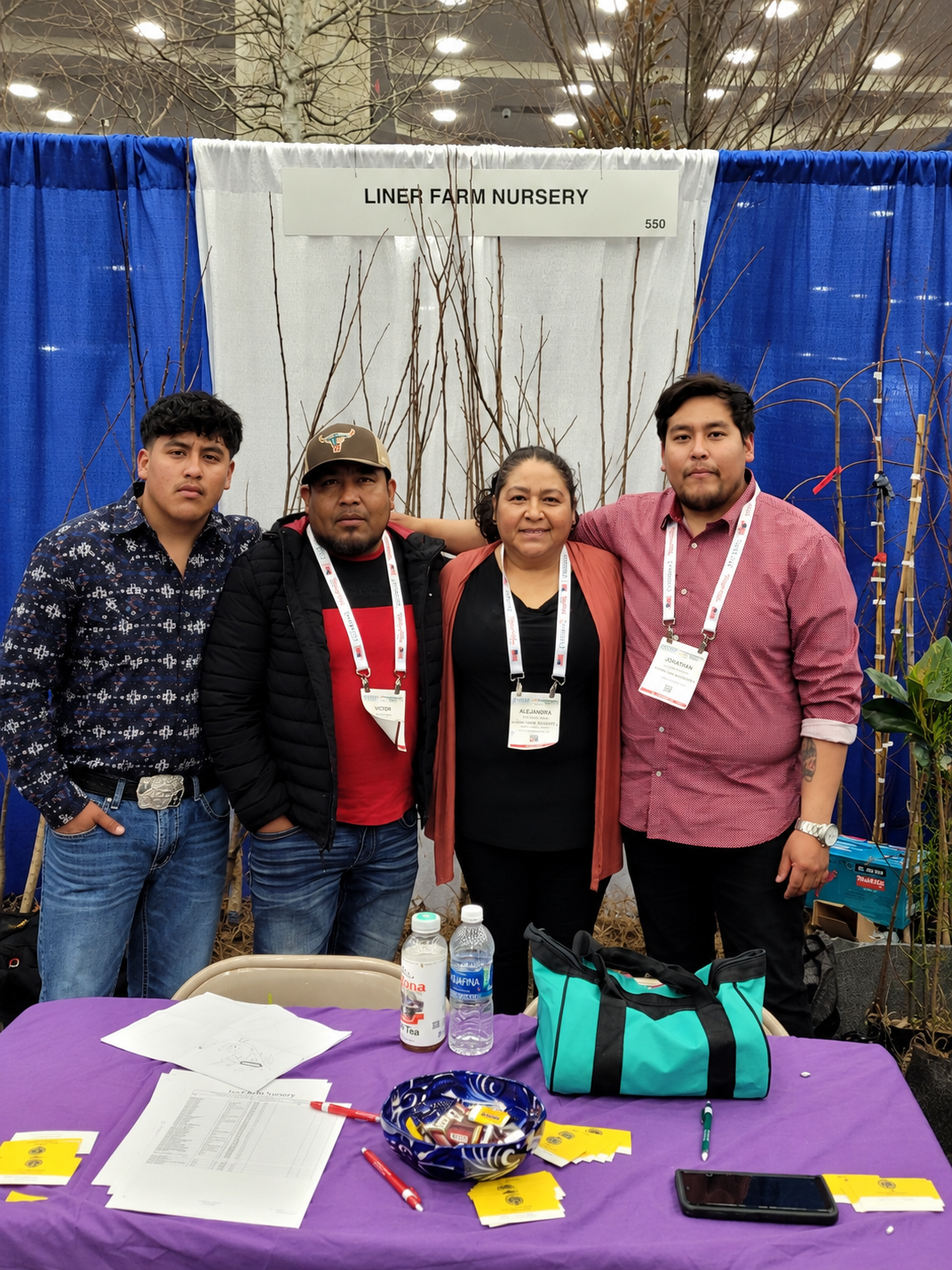Group of four people standing in front of a booth at a nursery exhibition, labeled "Liner Farm Nursery," with a table in front holding papers, pens, bottled water, and small yellow cards.