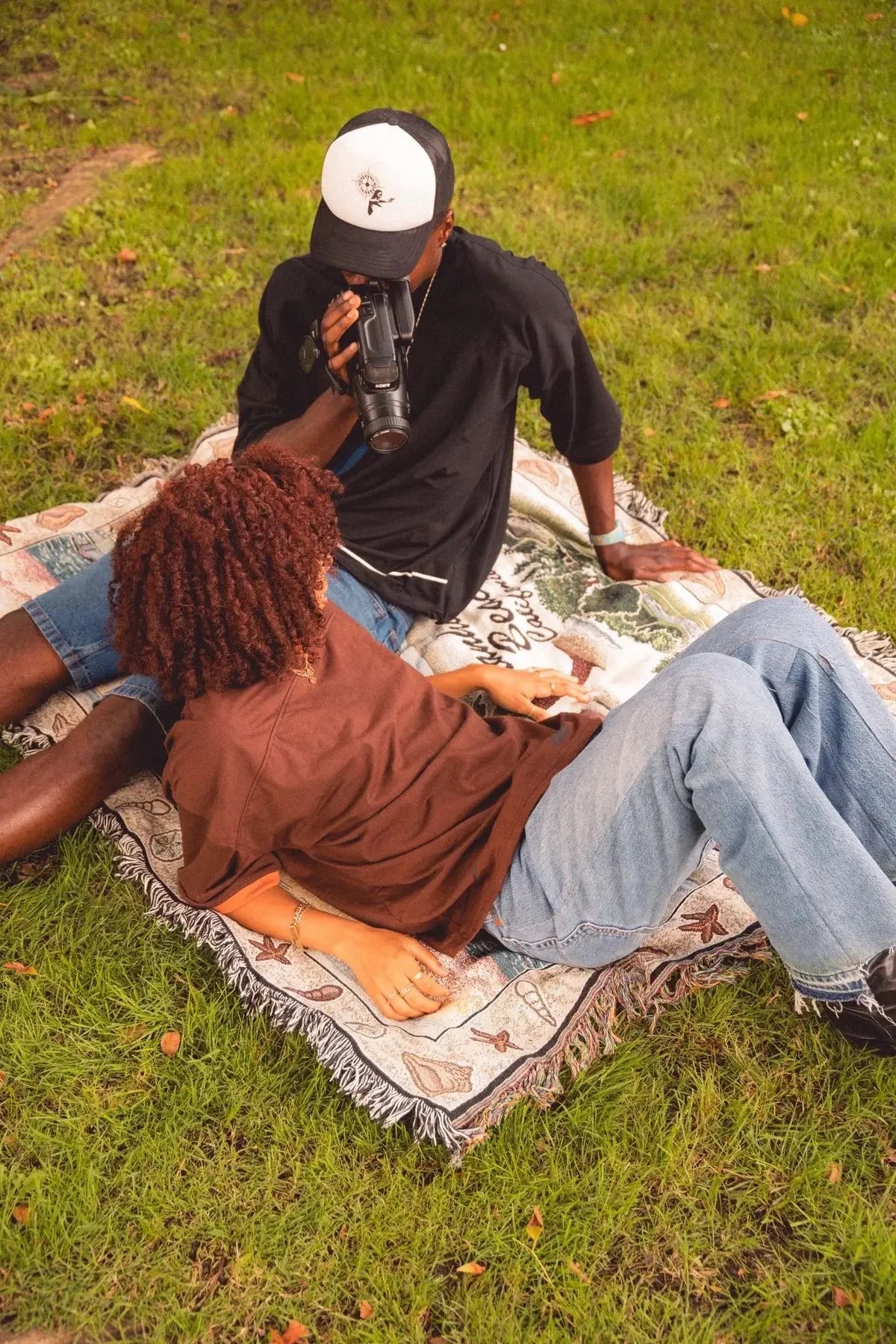 Two people on a blanket in a grassy outdoor setting, one person lying down and the other sitting, with a video camera.