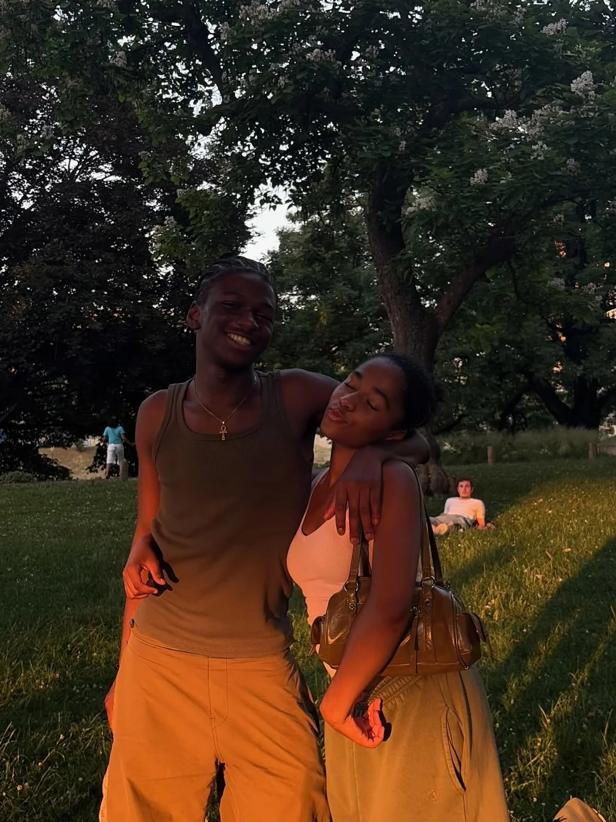 Two women, one with dark hair and the other with lighter hair, standing close together outdoors under a tree, smiling and embracing. In the background, a person is lying on the grass and another is walking.