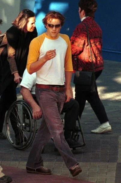 A person with reddish hair wearing sunglasses, a white t-shirt with yellow sleeves, and brown pants, pushing a wheelchair in an outdoor urban area.