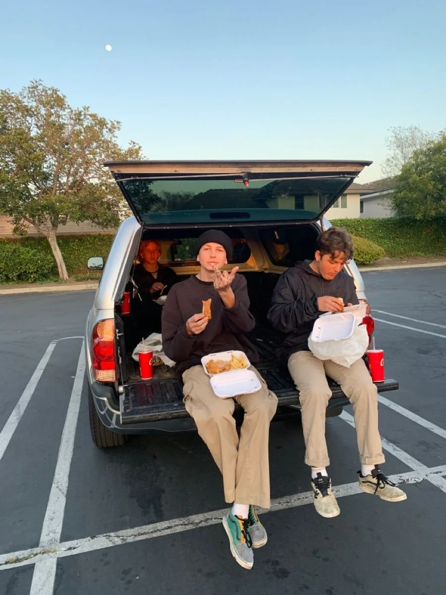 Three young people sitting in the open trunk of a silver SUV eating fast food in a parking lot during the daytime.