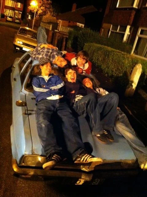 Four young people sitting and lying on the hood of a small silver car parked on a street at night, smiling and laughing.