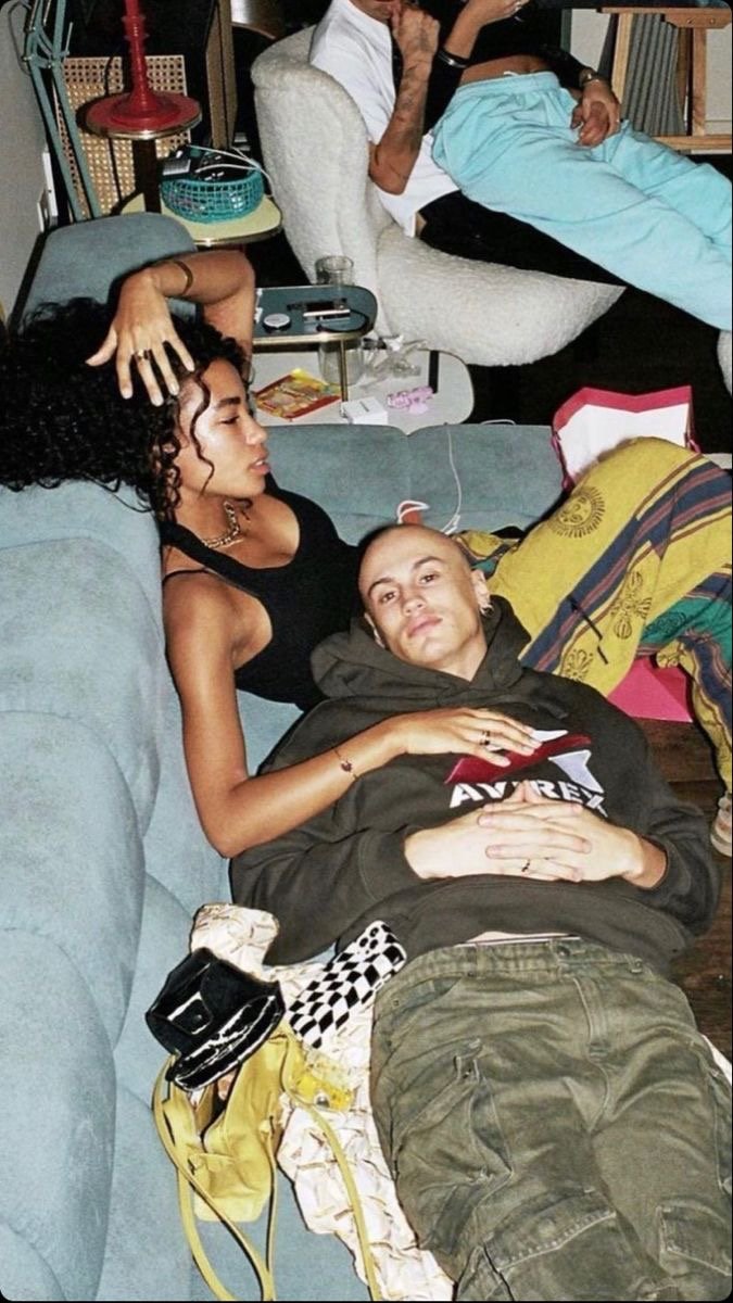 Two people are lying on a couch, one with curly hair and a black top, the other with short hair and a dark hoodie. They are resting with one person's hand on the other's head, and the second person's hand on their stomach. There are various objects a